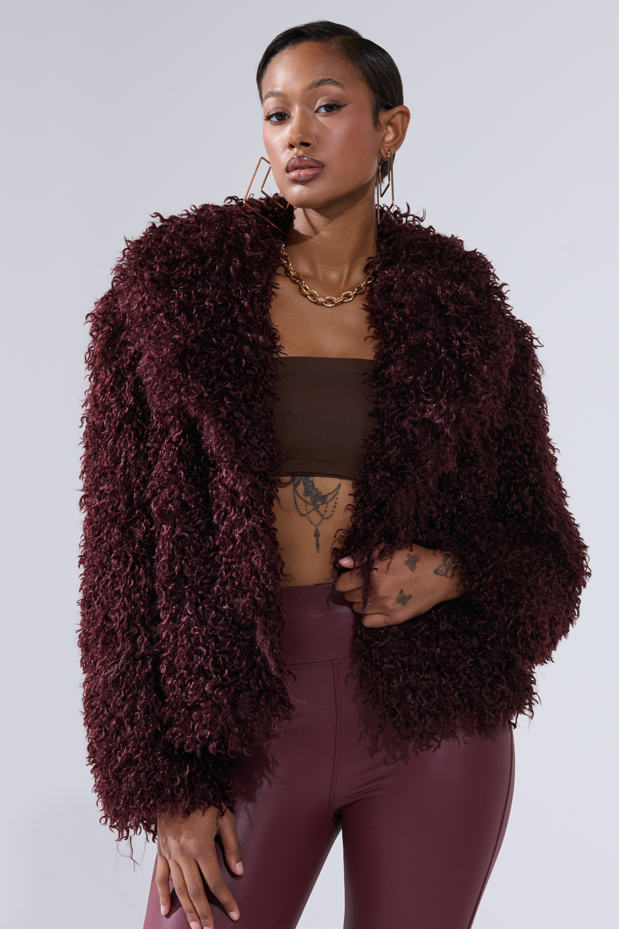 A woman stands confidently against a light gray background, wearing the VENUS FAUX FUR COAT—a curly, burgundy statement piece—paired with maroon high-waisted pants, a brown tube top, gold hoop earrings, and a chunky gold chain necklace.