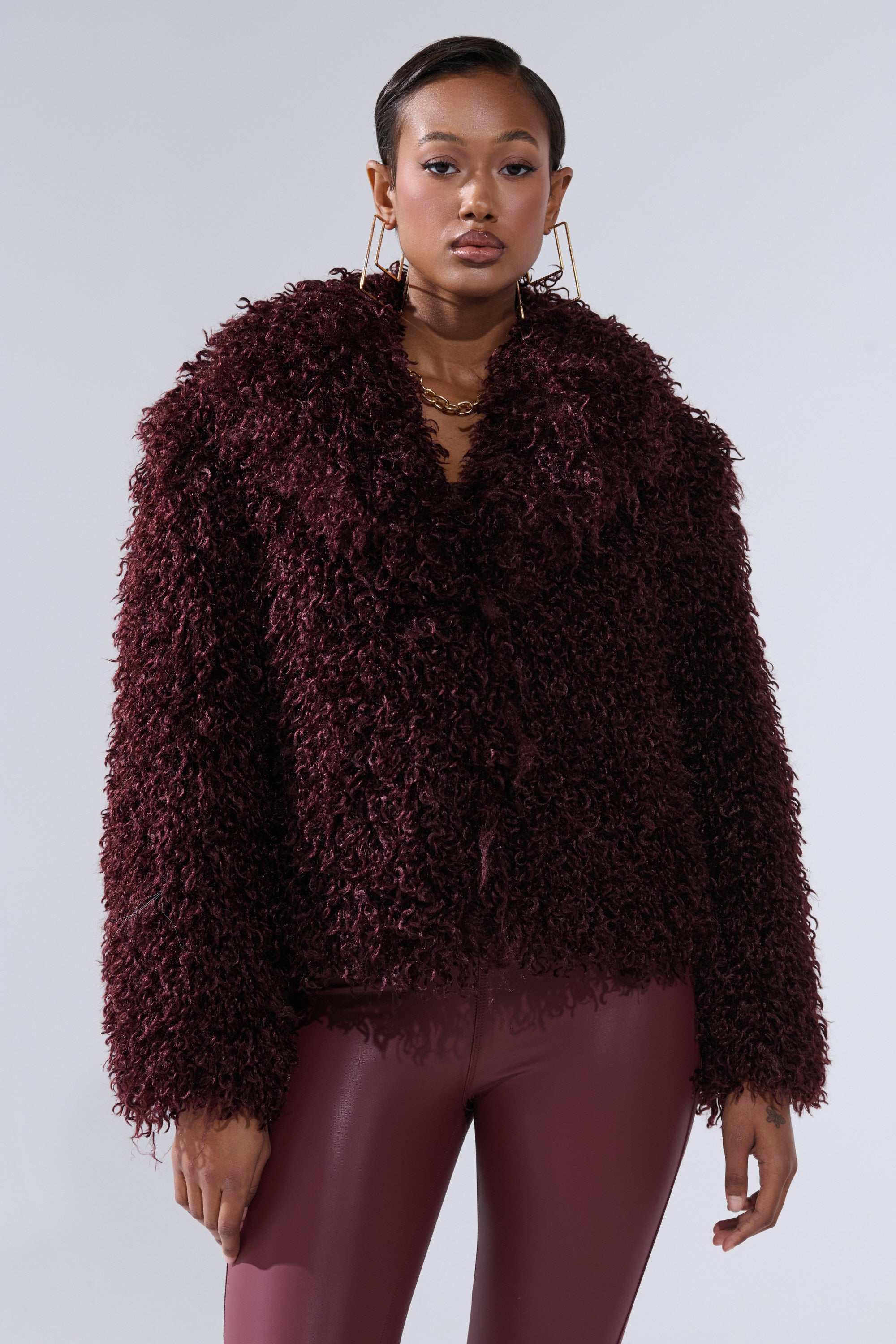 A woman in a burgundy VENUS FAUX FUR COAT with matching shiny high-waisted pants poses against a plain light background, her hair slicked back and large hoop earrings completing the look.