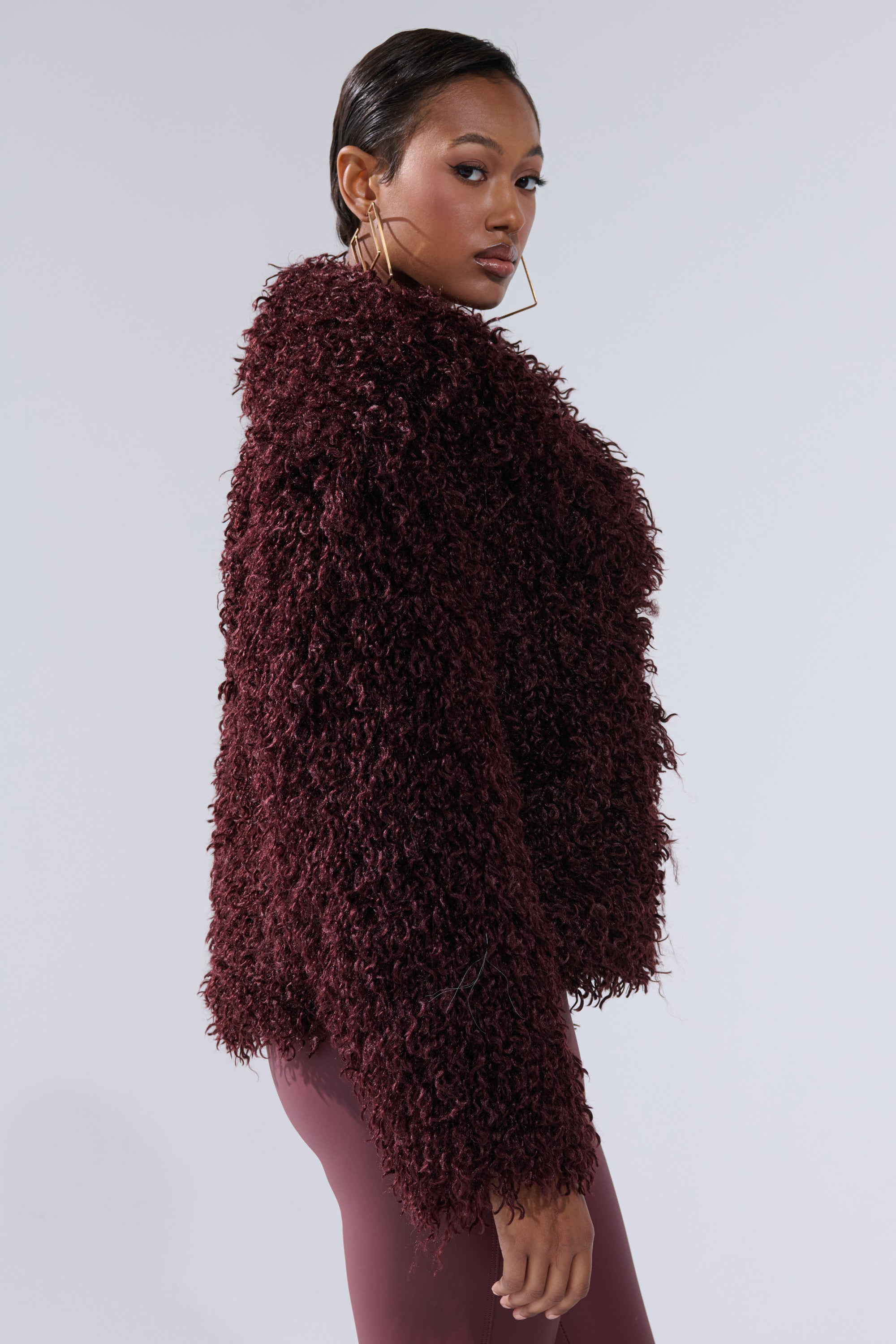 A woman in large hoop earrings and the VENUS FAUX FUR COAT from AKIRA Label gazes over her shoulder against a plain light gray background.