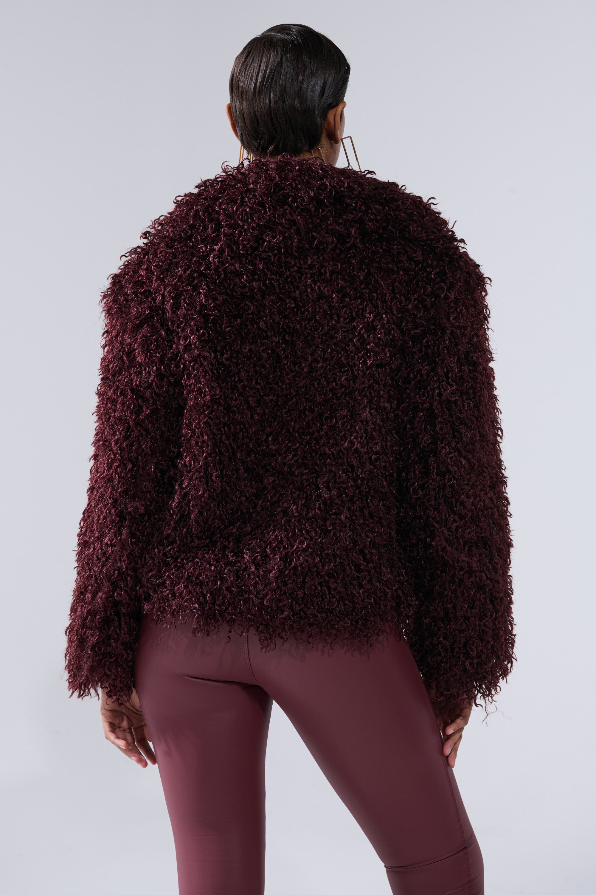 A person with short dark hair, seen from behind, wears the VENUS FAUX FUR COAT with matching burgundy fitted pants against a plain light gray background.