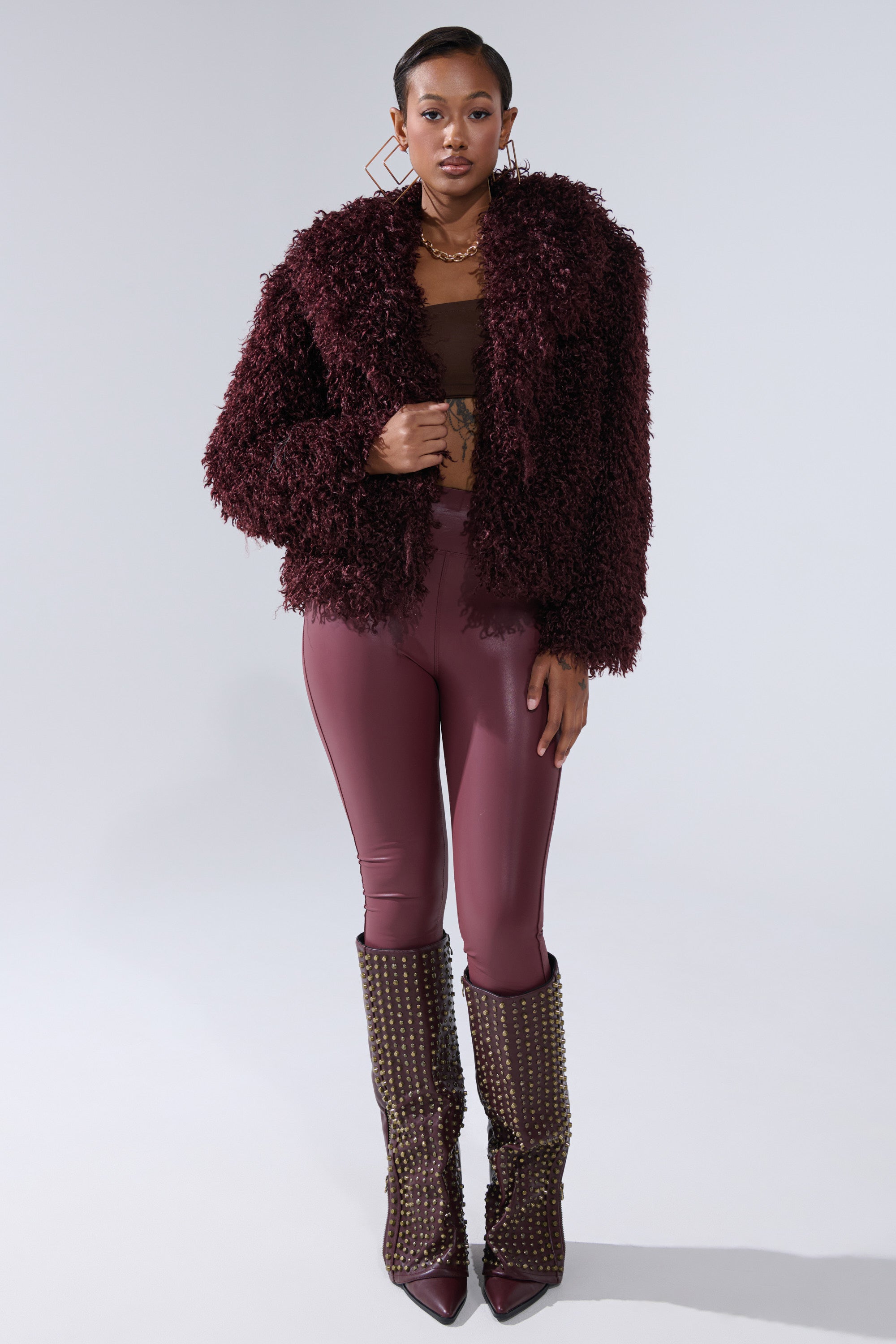 A woman with short hair and gold jewelry poses against a plain light background, wearing the VENUS FAUX FUR COAT, burgundy faux-leather leggings, a brown mesh top, and tall studded boots.