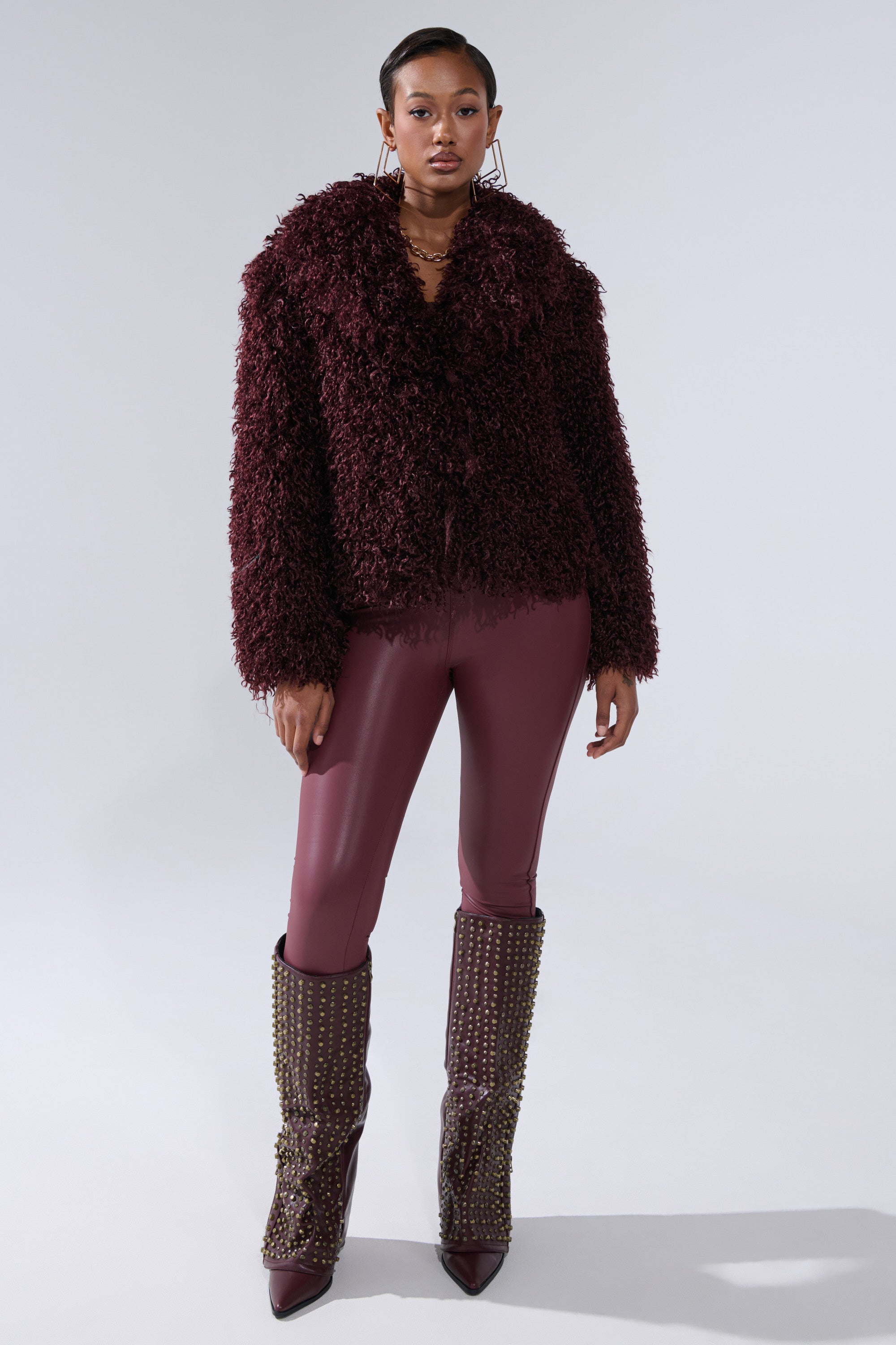 A woman stands facing forward in the VENUS FAUX FUR COAT, featuring a shaggy burgundy texture, paired with shiny burgundy leggings and tall studded burgundy boots, set against a plain light gray background.