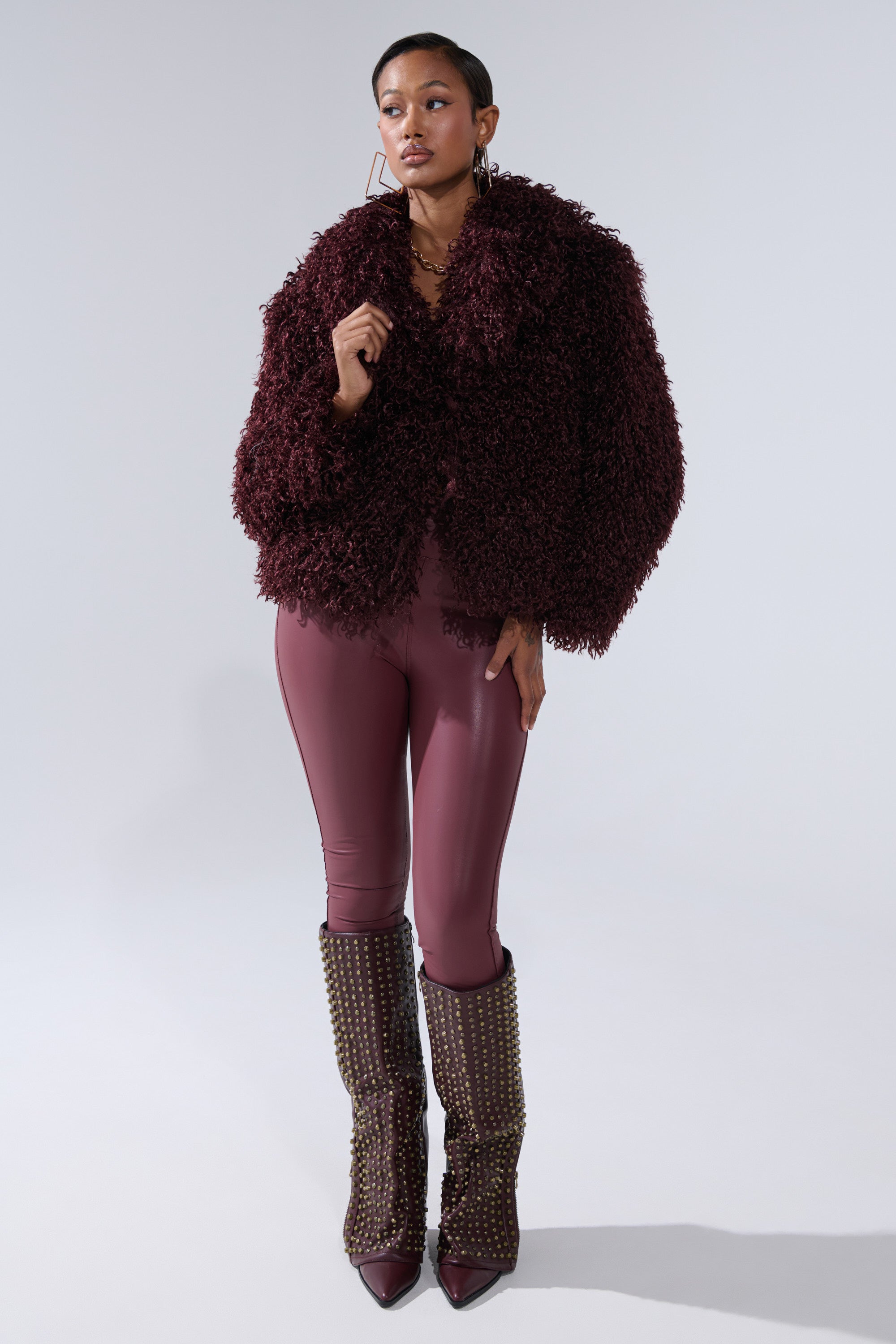 A woman models the VENUS FAUX FUR COAT in burgundy with a shaggy texture, paired with matching shiny burgundy leggings and knee-high pointed studded boots, set against a plain light background.