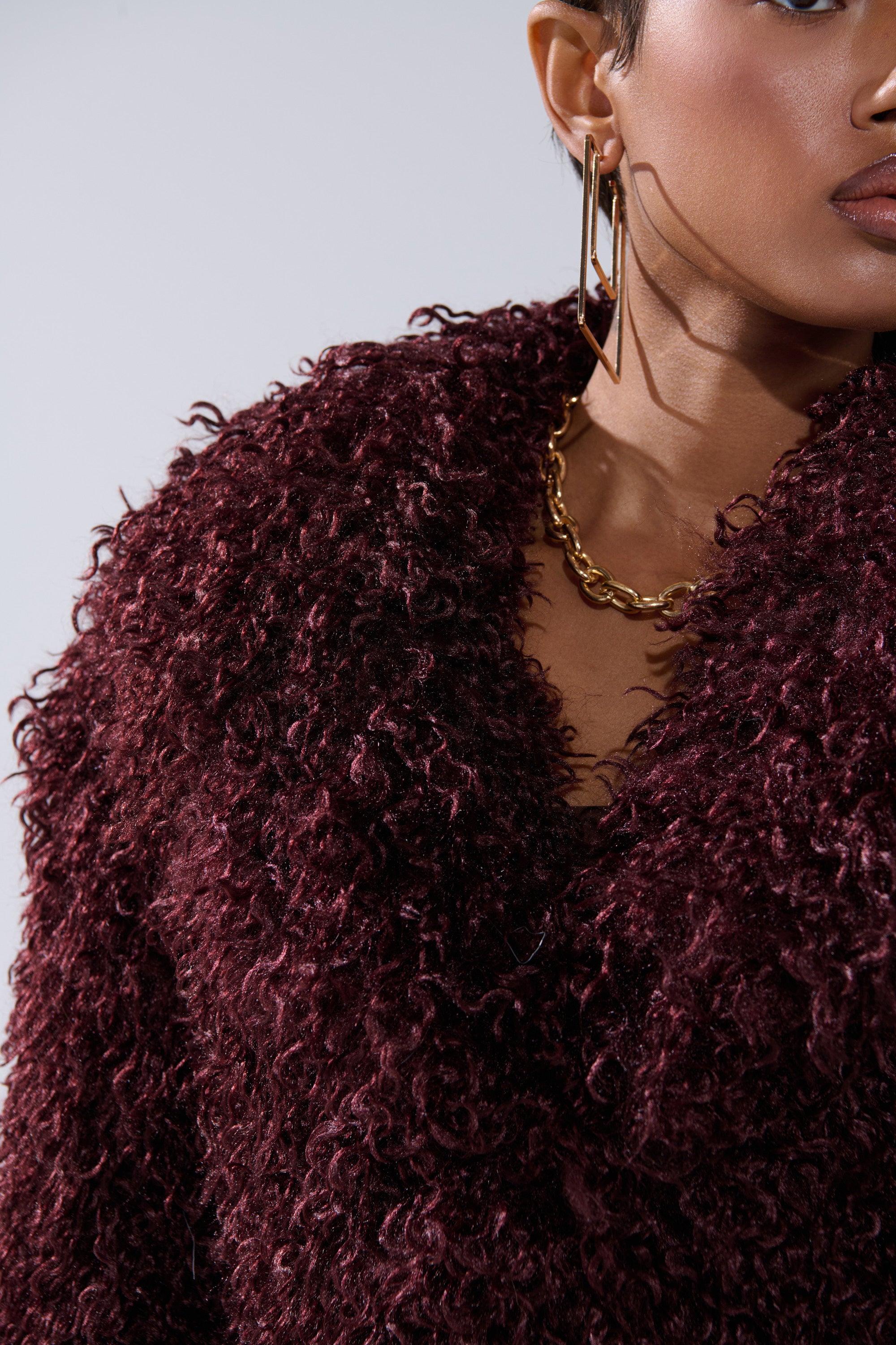 A close-up shows the lower face and upper body of a person wearing the VENUS FAUX FUR COAT in deep burgundy, styled with a chunky gold chain necklace and large gold hoop earrings. The coat features a shaggy faux fur texture.