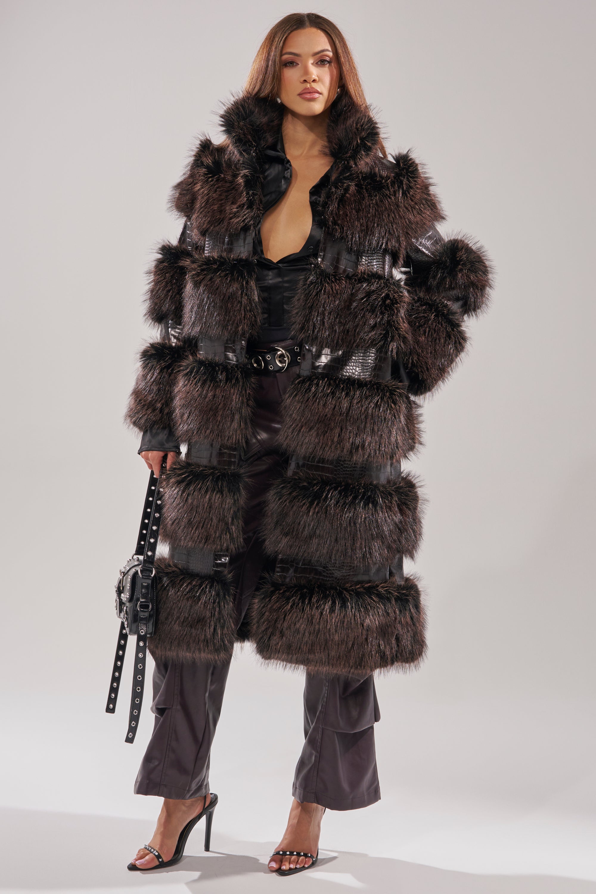 A woman stands confidently in the MOXIE FUR TRIM TRENCH with leather accents, paired with black belted pants, high-heeled sandals, and a black handbag against a neutral background.