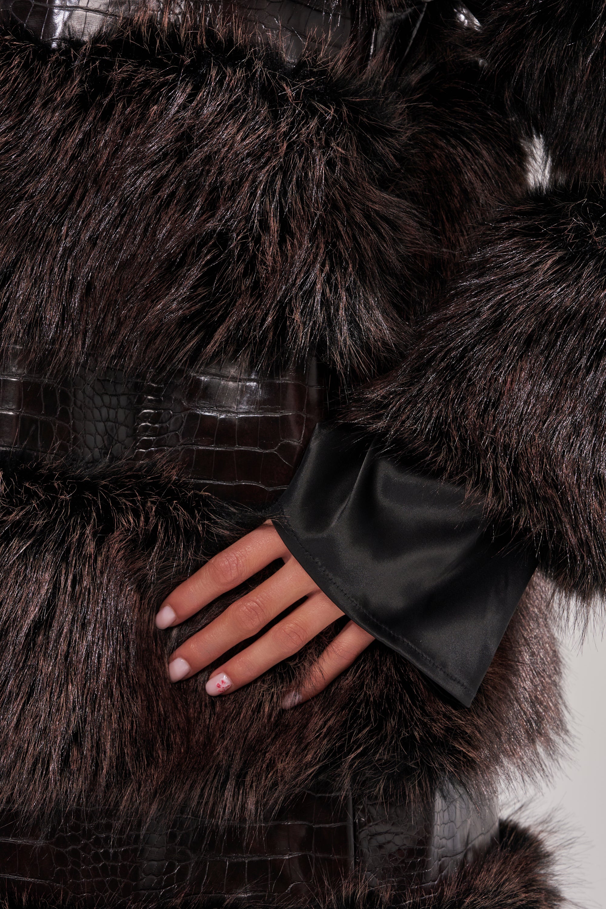 A hand with light pink nails and red designs rests on the MOXIE FUR TRIM TRENCH, showcasing its dark brown faux fur texture and black crocodile-patterned leather.