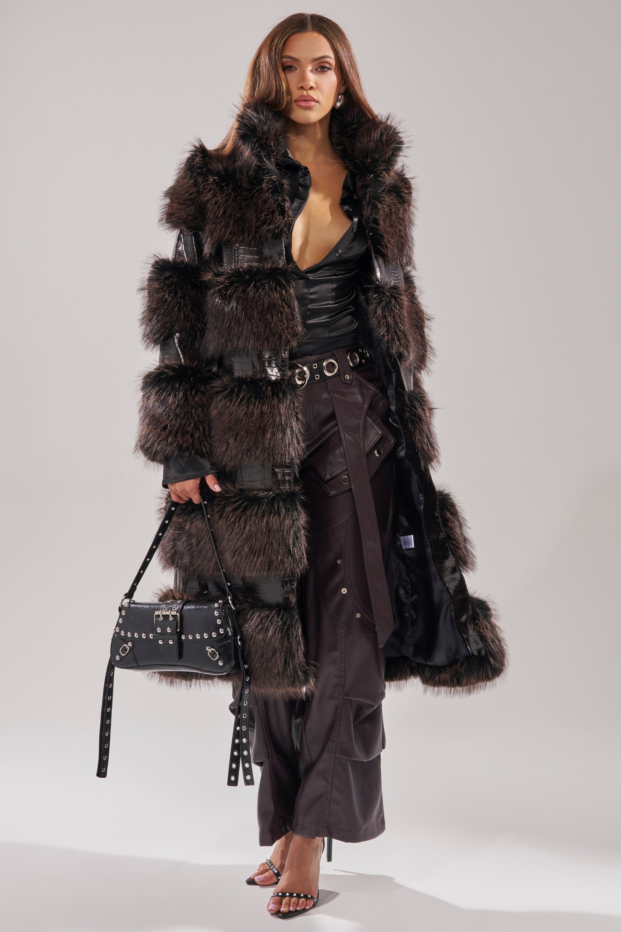 Wearing the MOXIE FUR TRIM TRENCH with dark faux fur and leather, a woman poses against a light grey background over a black V-neck top, high-waisted pants, studded black handbag, and heeled sandals.