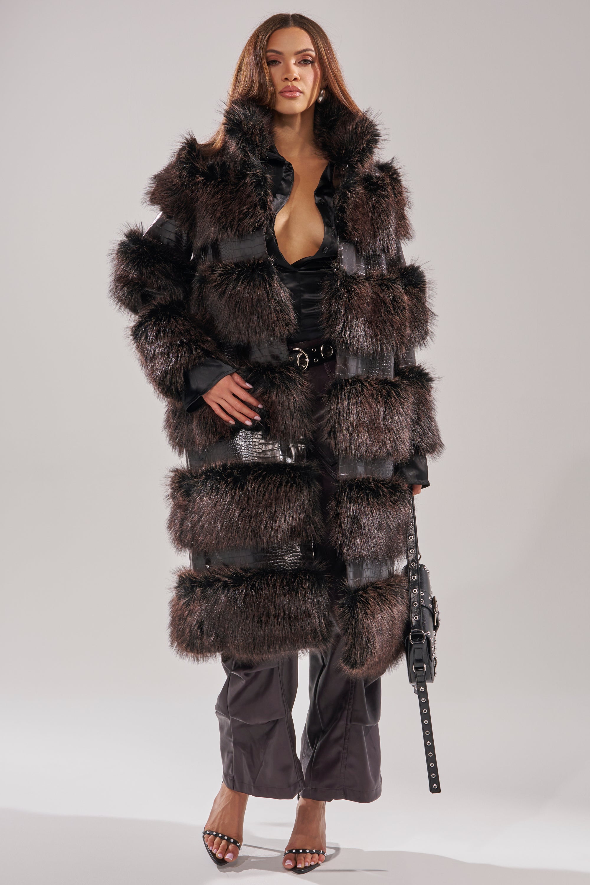 A woman stands against a plain background wearing the MOXIE FUR TRIM TRENCH in brown and black over a black outfit with wide-legged pants, strappy high heels, and holding a black handbag at her side.