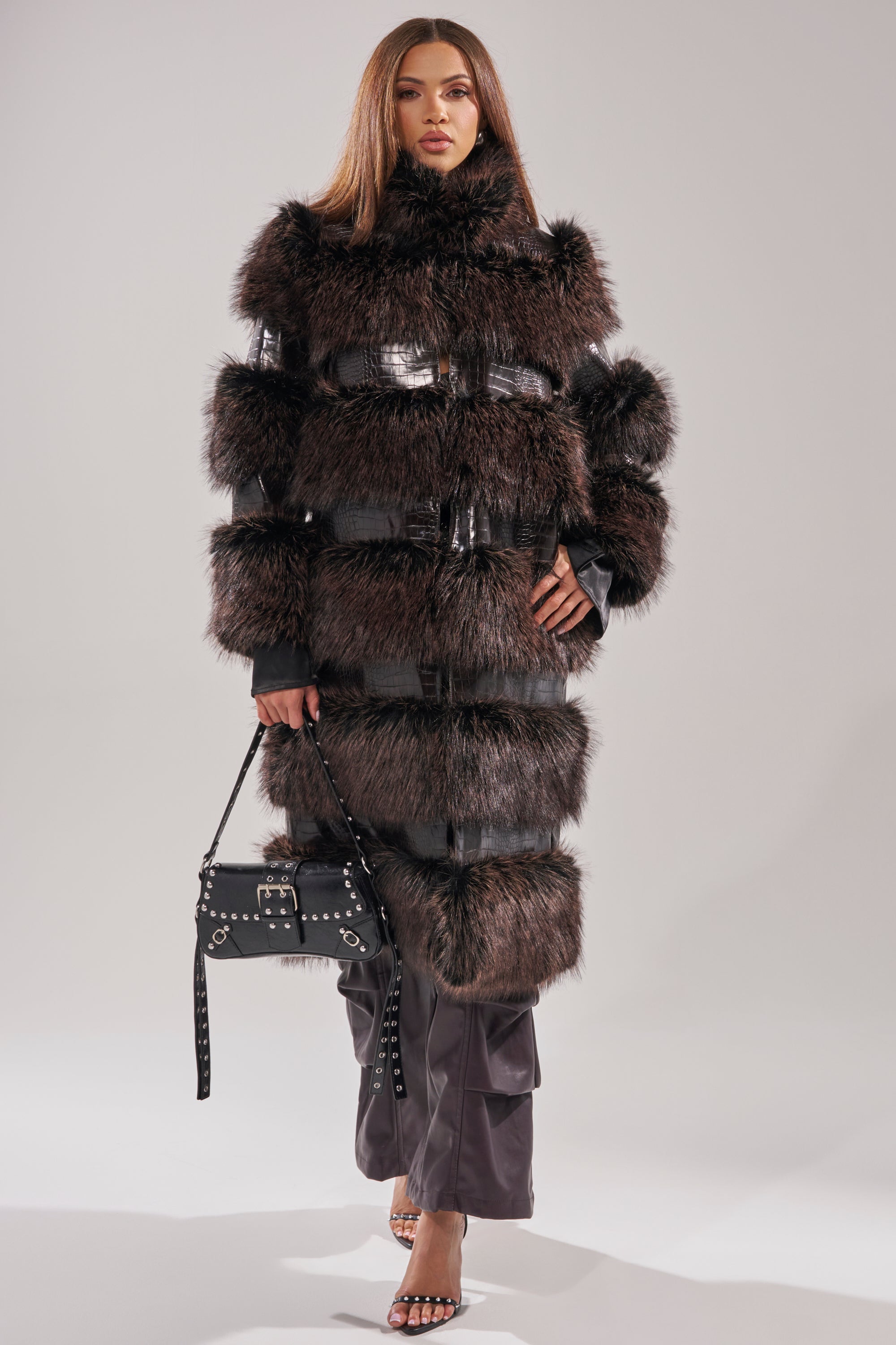 A woman wears the MOXIE FUR TRIM TRENCH, a longline dark brown faux fur coat with horizontal leather bands, paired with a studded black handbag, heeled sandals, and leather pants against a plain light background.