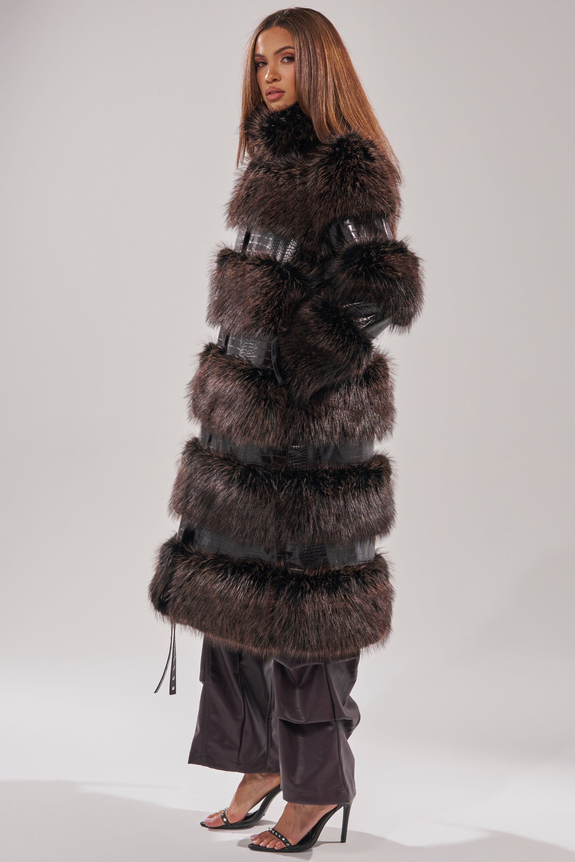 A woman stands in a studio wearing the MOXIE FUR TRIM TRENCH, dark wide-leg pants, black high-heeled sandals, and with straight brown hair. She faces slightly to the left against a plain background.