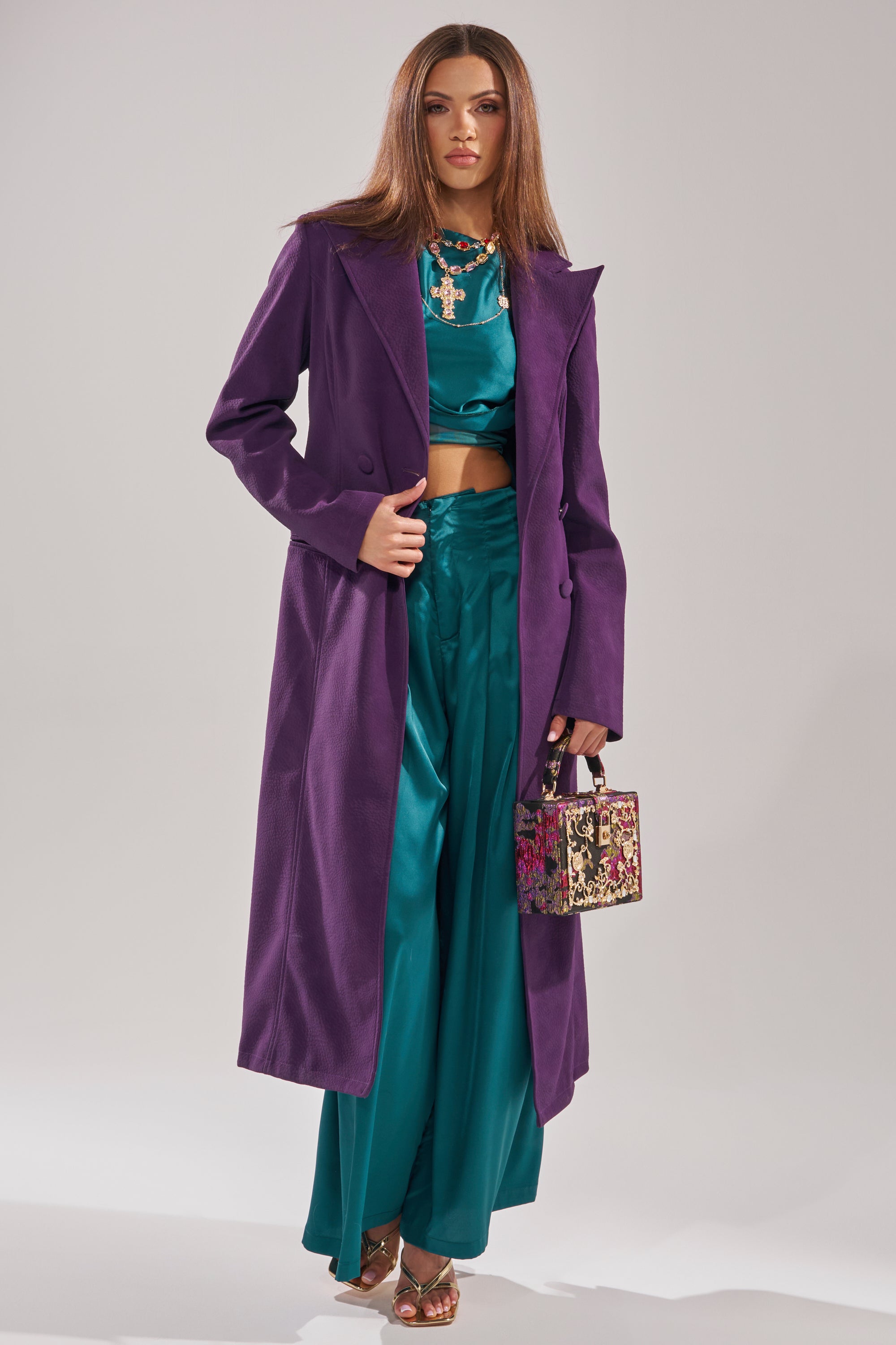 A woman wears the MAEVE SUEDE FEEL TRENCH in purple over a teal satin crop top and wide-leg pants. She accessorizes with a small patterned handbag, gold heels, straight hair, and bold makeup.