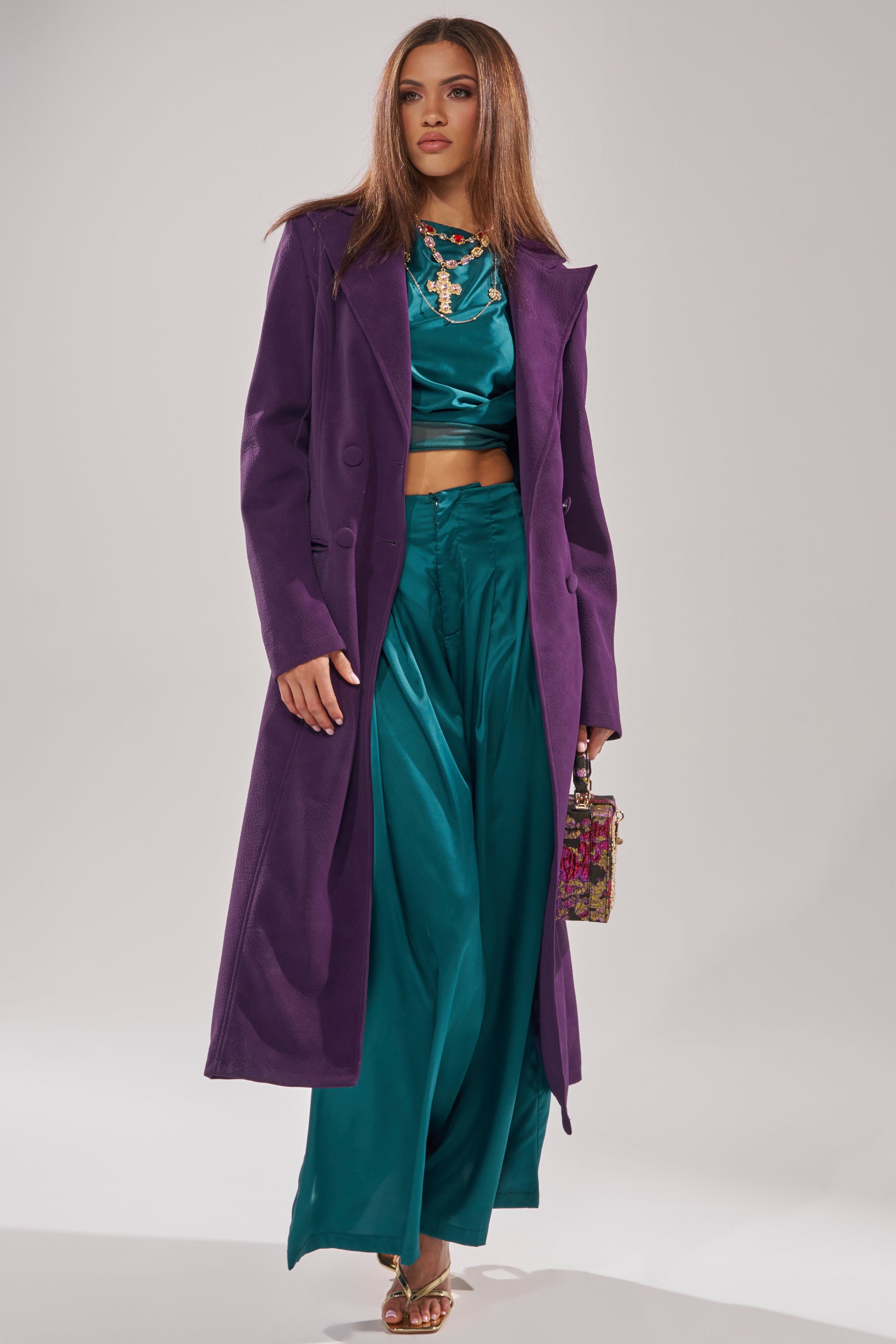 A woman with straight brown hair and a confident expression wears the MAEVE SUEDE FEEL TRENCH in purple over a shiny teal top and matching wide-leg pants, accessorized with a small patterned handbag and gold sandals.