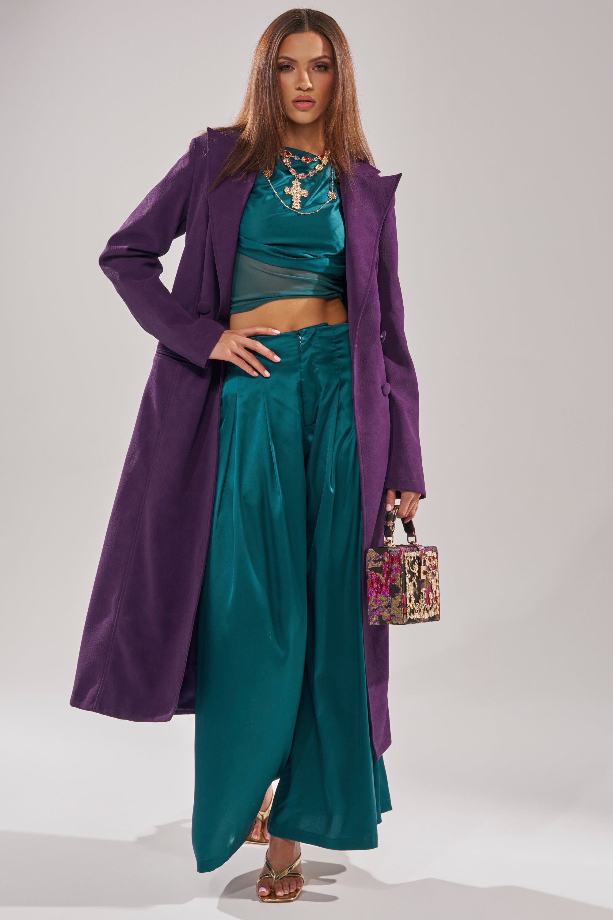 A woman poses confidently against a plain background wearing the MAEVE SUEDE FEEL TRENCH over a teal crop top and wide-leg pants, styled with layered necklaces, heeled sandals, and a small floral handbag.