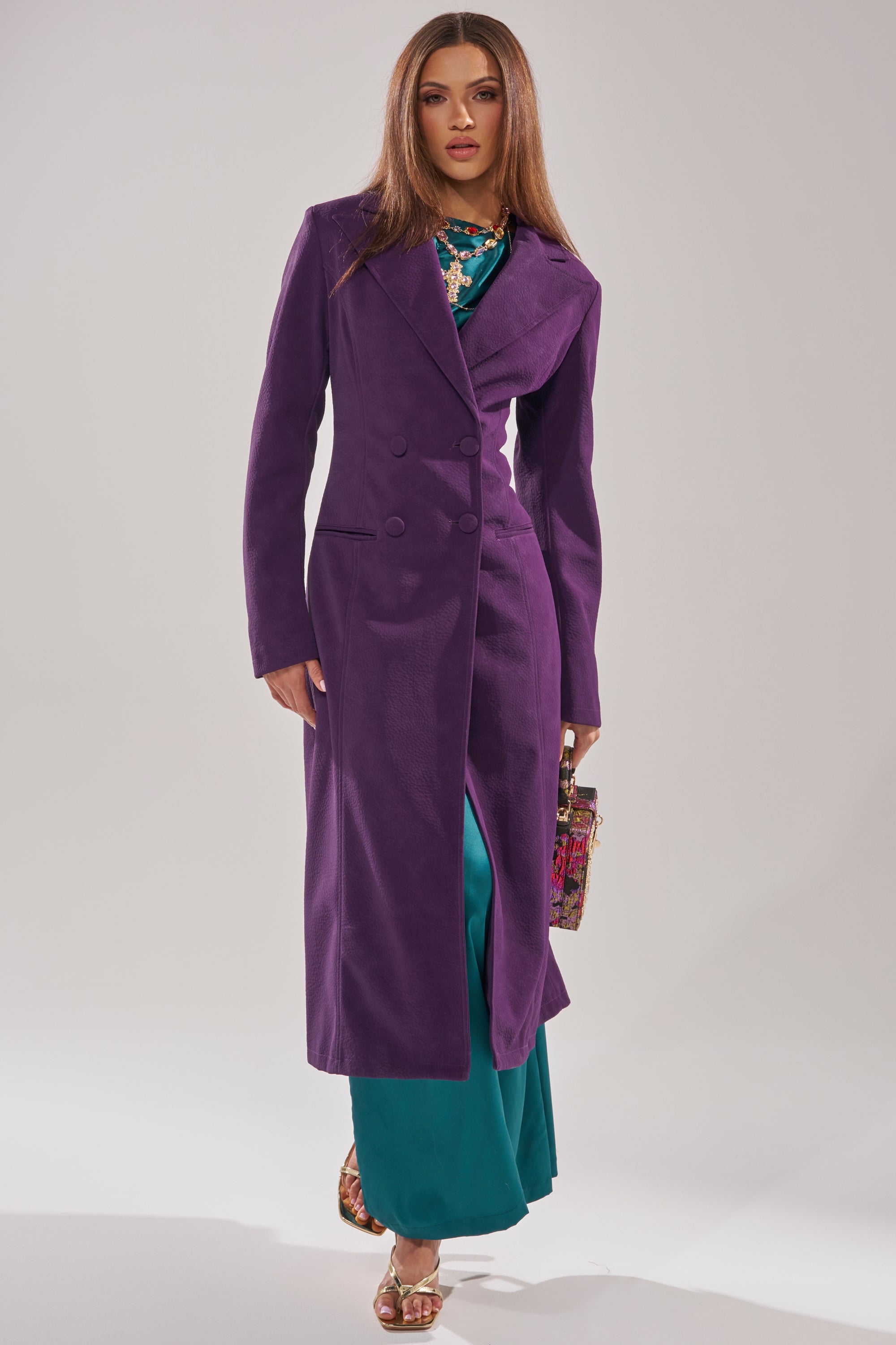 A woman wearing the MAEVE SUEDE FEEL TRENCH, a longline double-breasted purple coat, over a teal dress with gold sandals and a colorful handbag stands against a plain light background.