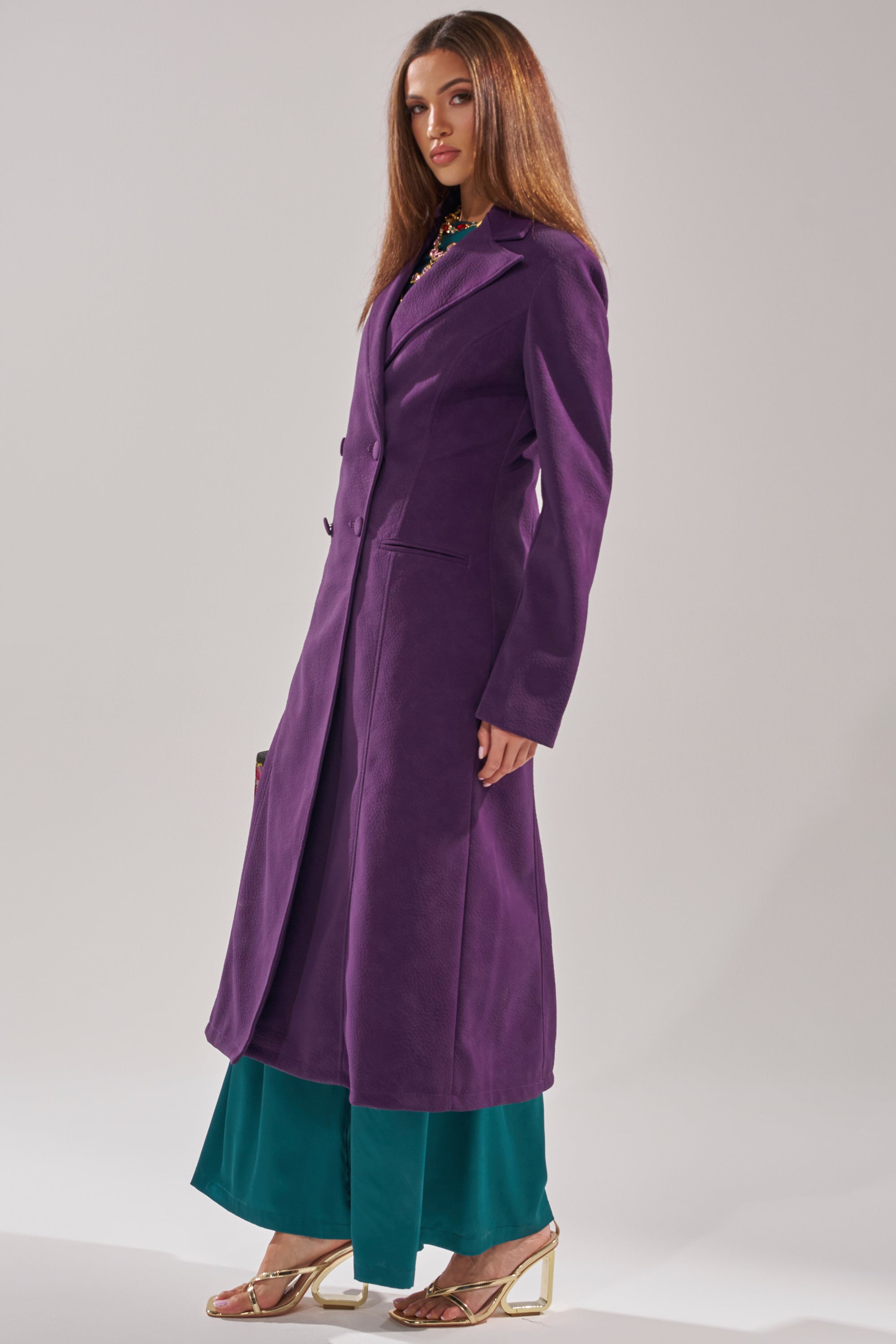 A woman stands confidently against a plain light background wearing the MAEVE SUEDE FEEL TRENCH in deep purple over teal wide-leg pants and gold high-heeled sandals, her straight brown hair framing her face.