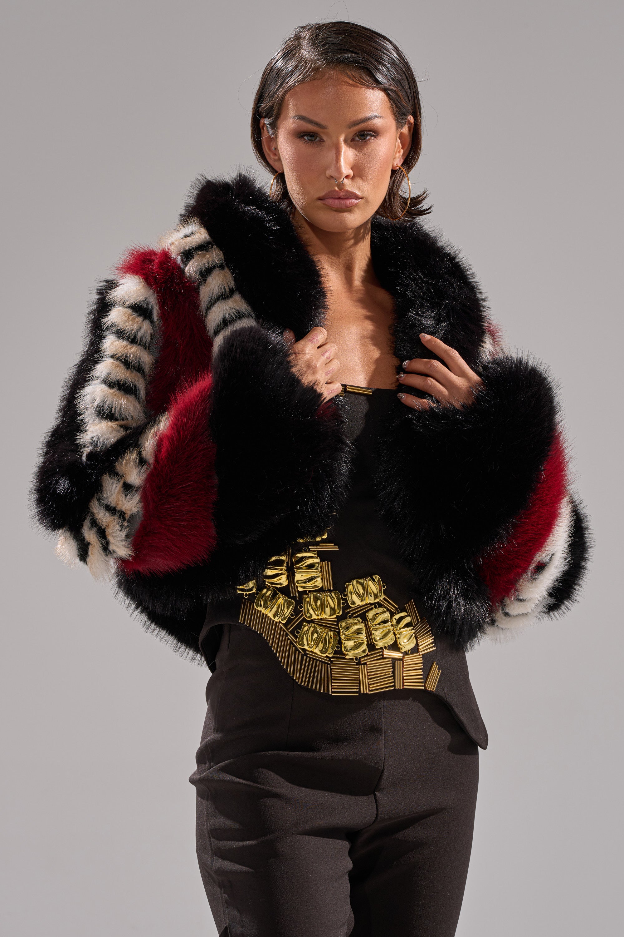A woman poses in the HARD TO FIND PANELED FAUX FUR, a bold color block cropped jacket with black, red, and white stripes, styled over a black top with gold embellishments and black pants against a plain light gray background.