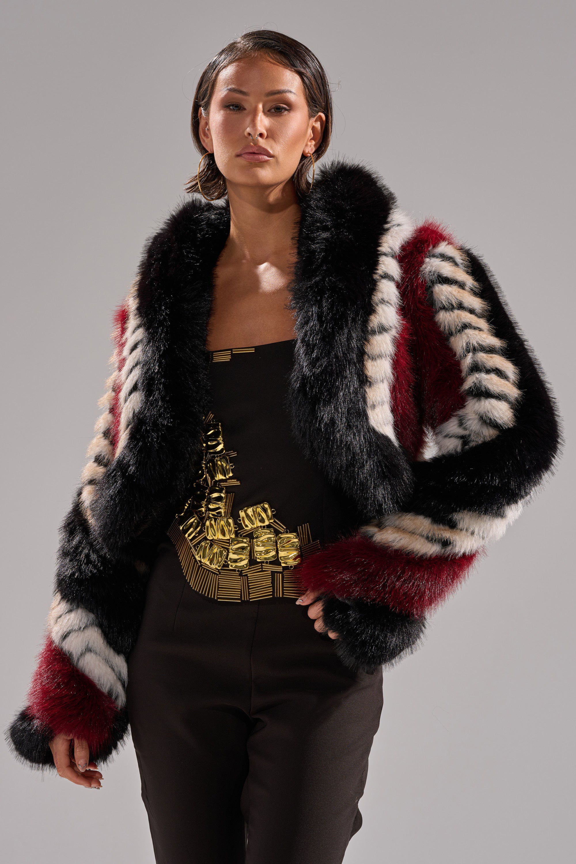 A woman confidently poses against a plain background wearing the HARD TO FIND PANELED FAUX FUR—a bold, cropped, boxy color block jacket in multicolored faux fur with black, white, and burgundy stripes—over a black outfit with intricate gold embellishment.