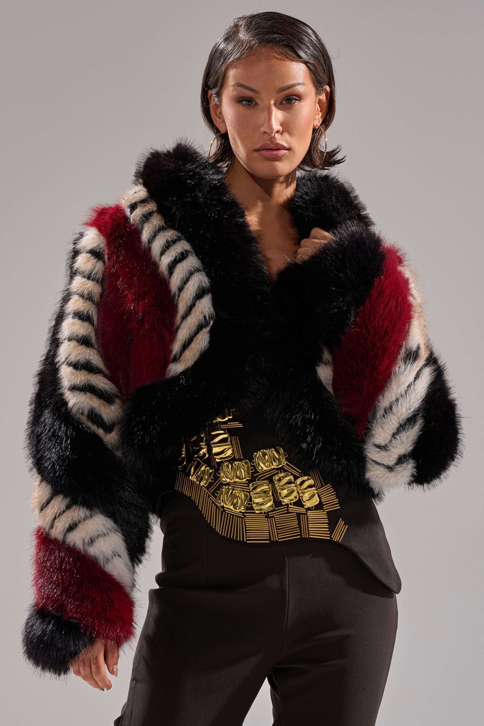A woman wears the HARD TO FIND PANELED FAUX FUR jacket featuring black, white, and red color block stripes over a black top with a gold geometric design, standing against a plain gray background.