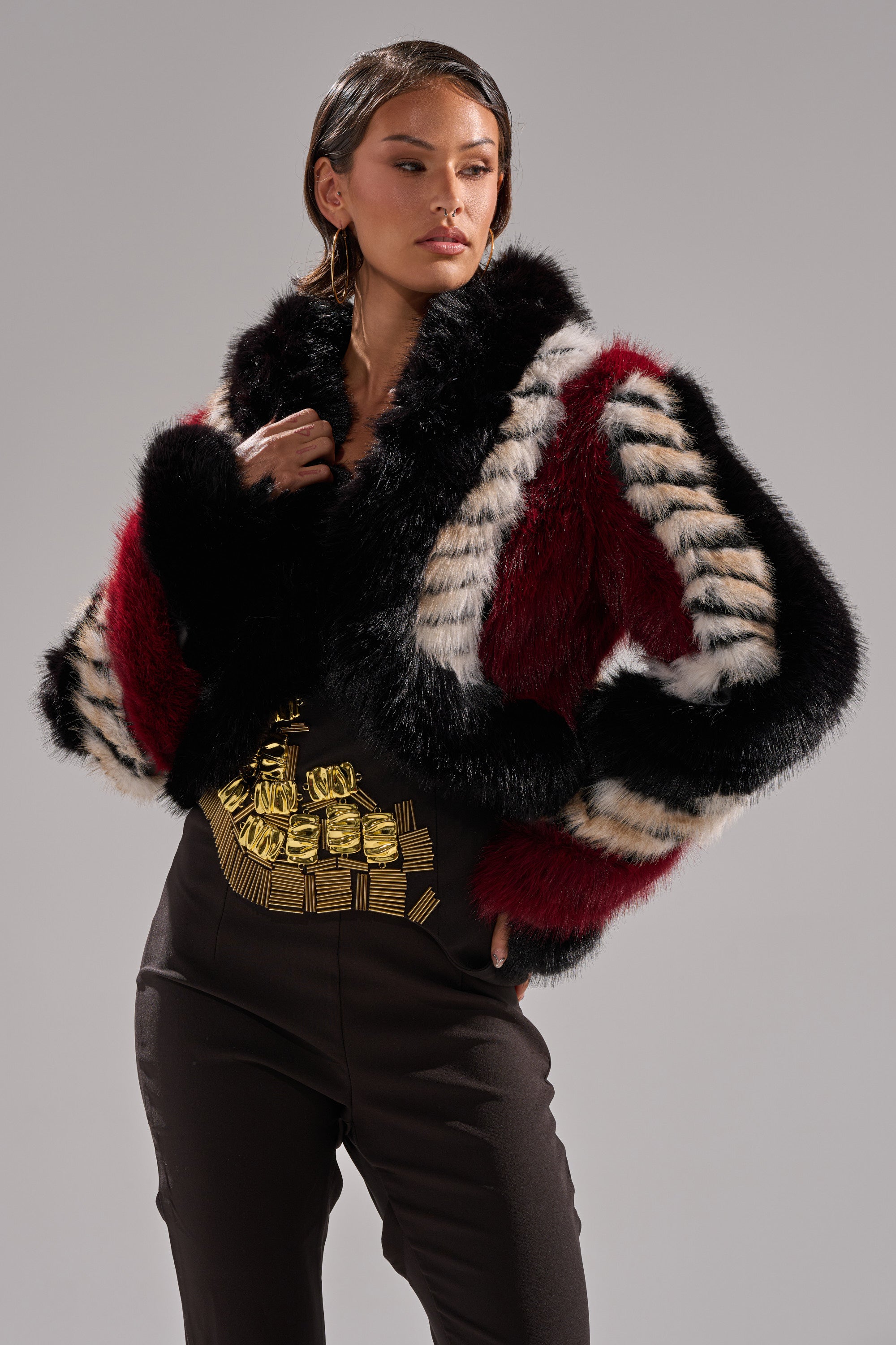 A woman models the HARD TO FIND PANELED FAUX FUR jacket with red, black, and white stripes, teamed with black pants featuring a bold gold belt motif, set against a plain gray background.