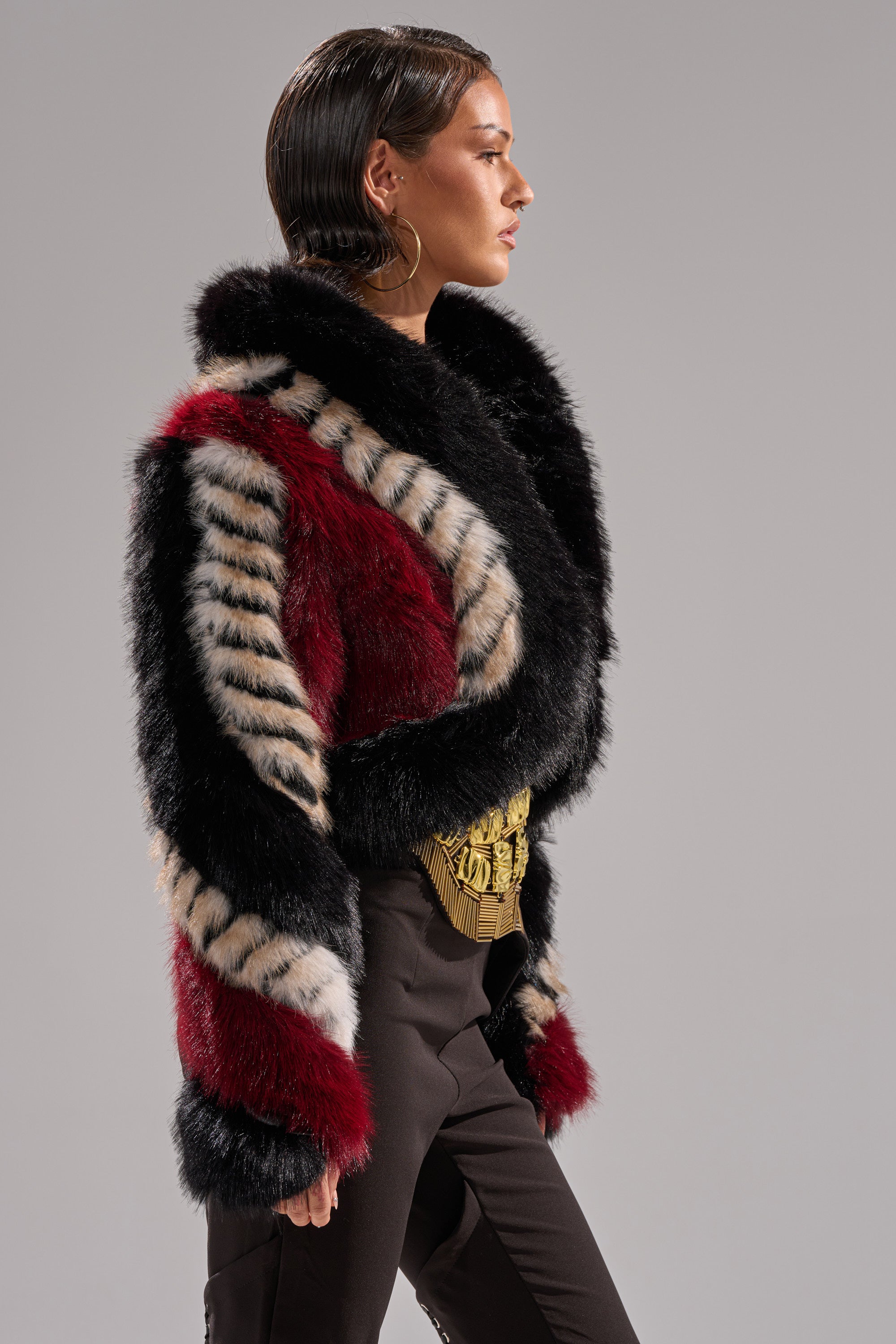 A woman with slicked-back hair models the HARD TO FIND PANELED FAUX FUR jacket featuring black, red, and beige panels, styled with high-waisted black pants and a gold belt, standing in profile against a plain gray background.