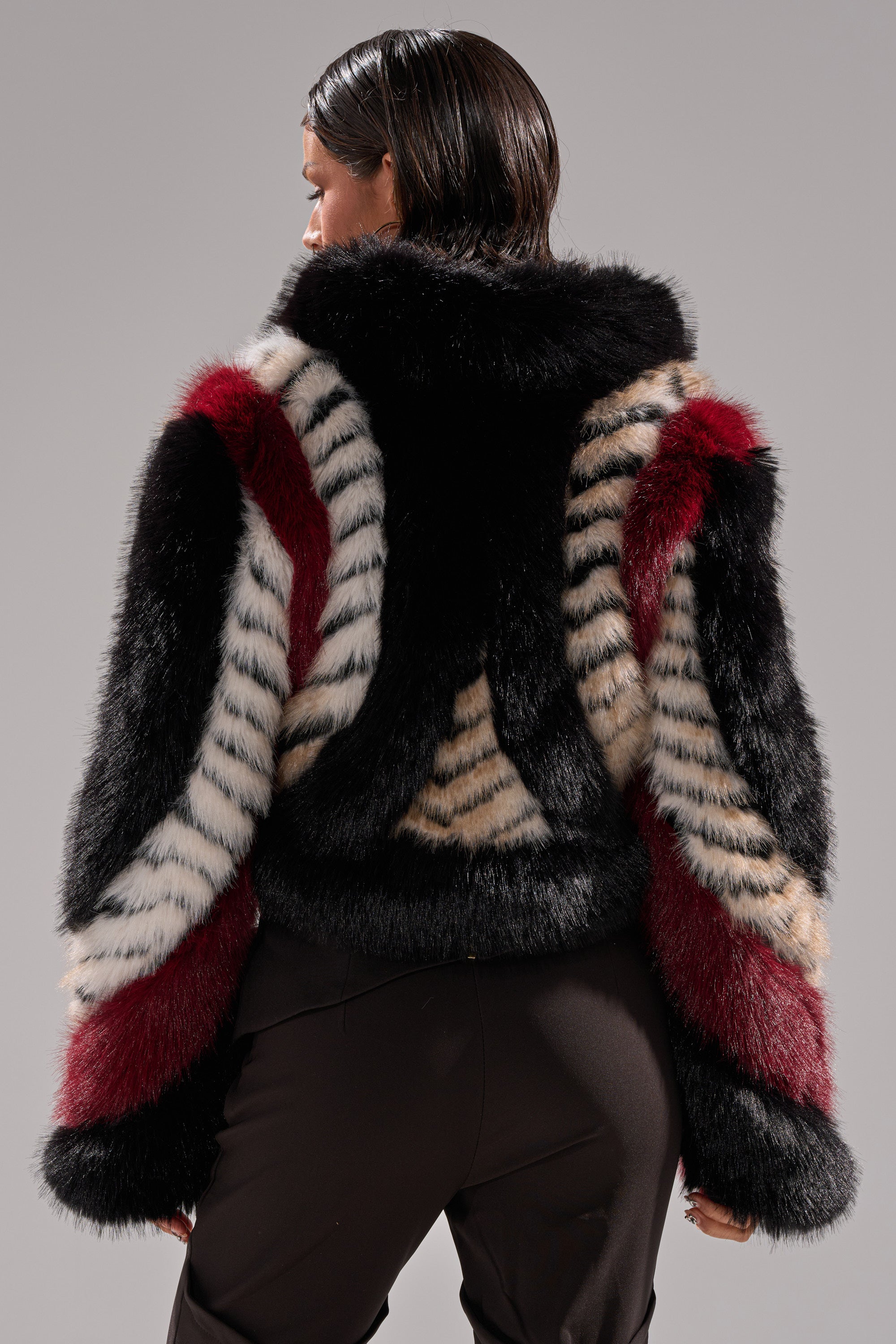 Someone with short dark hair is seen from behind wearing the HARD TO FIND PANELED FAUX FUR jacket, featuring bold black, red, ivory, and beige stripes, along with black pants against a neutral backdrop.
