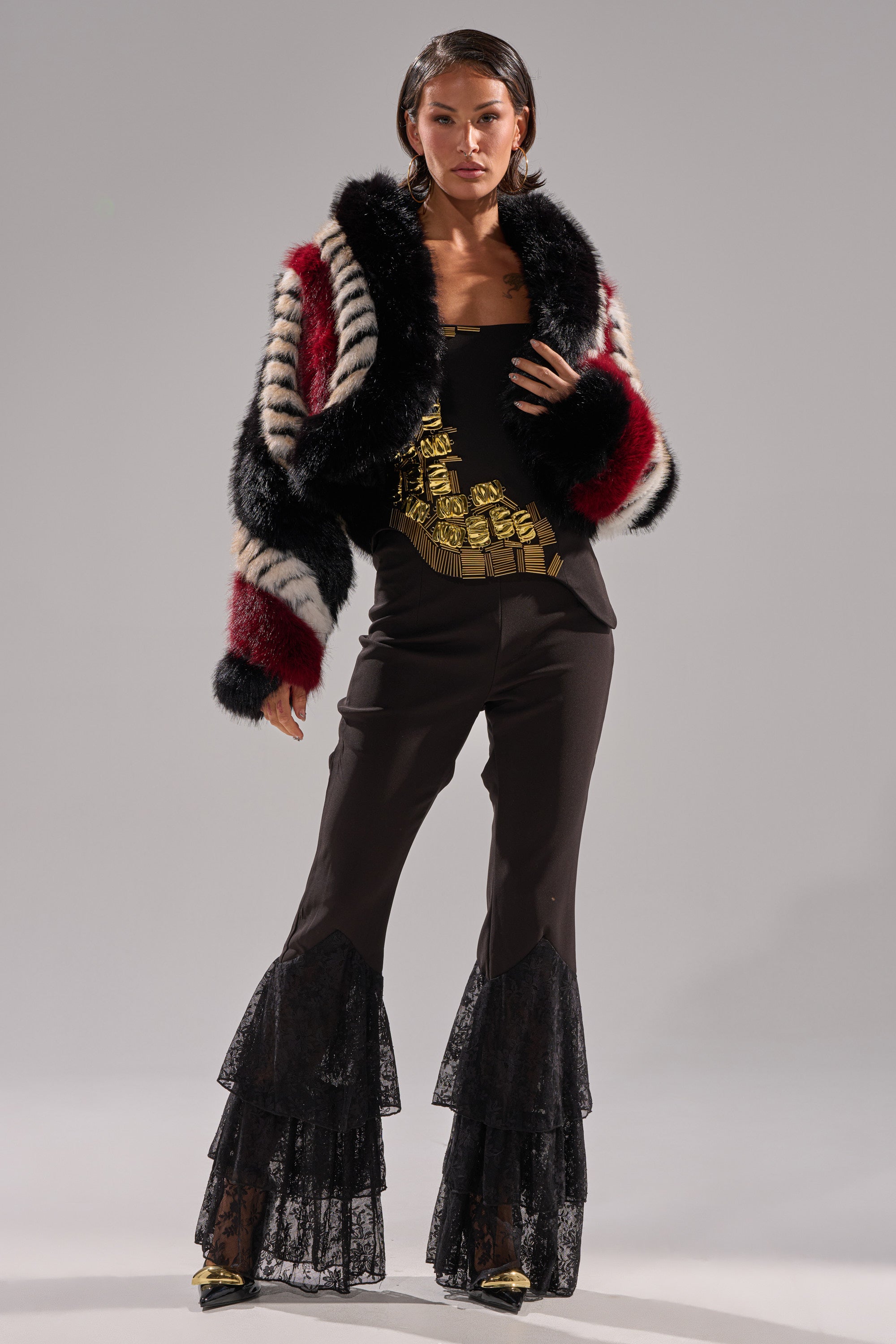 A woman wears the HARD TO FIND PANELED FAUX FUR jacket—cropped, boxy fit with black, white, and red stripes—paired with a black and gold top and layered black lace flared pants, posing against a plain gray background.