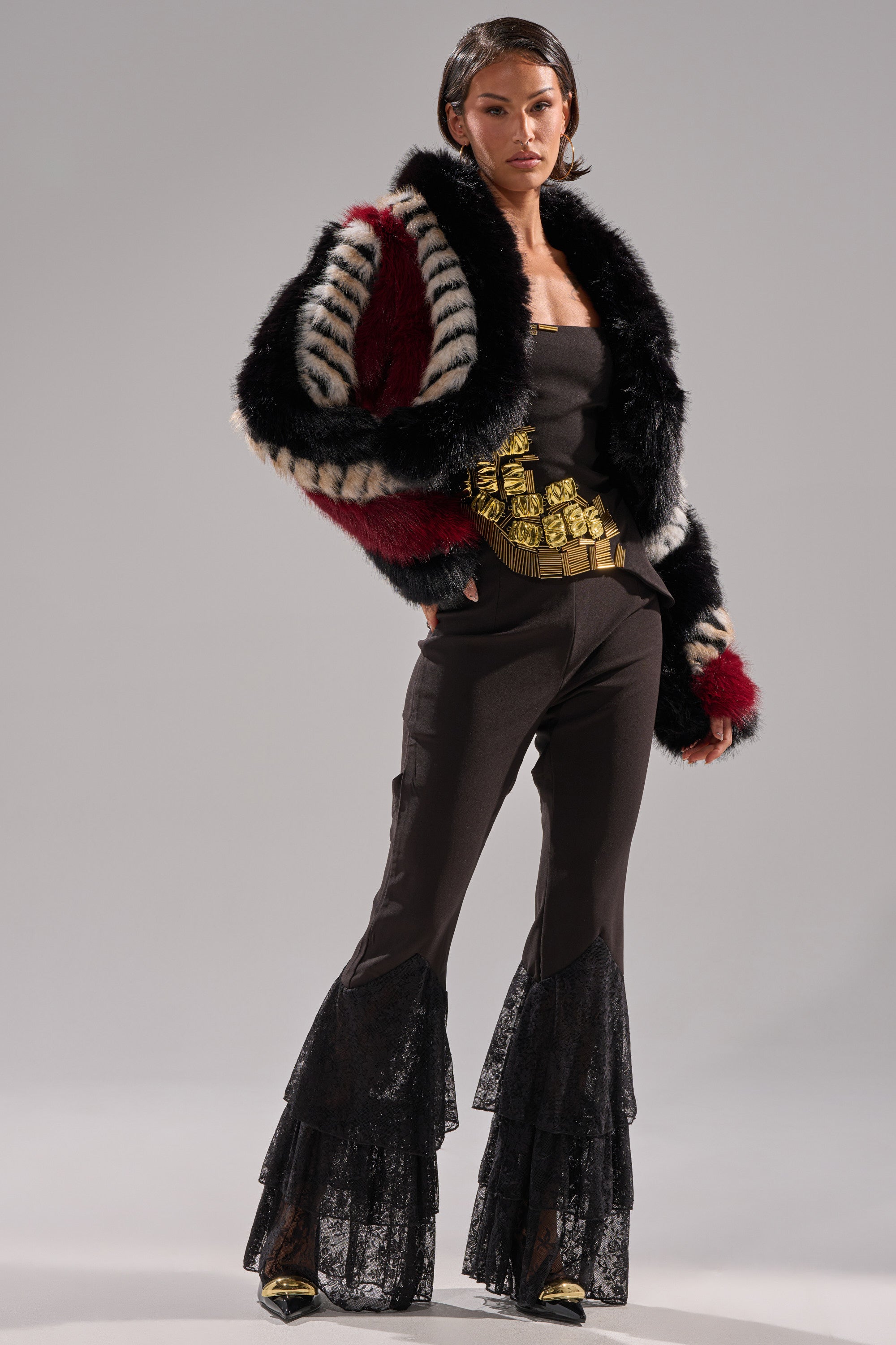 A person stands confidently in the HARD TO FIND PANELED FAUX FUR jacket with bold black, white, and red patterns, paired with a black strapless top, flared lace pants, gold belt, and black pointed shoes against a plain gray background.