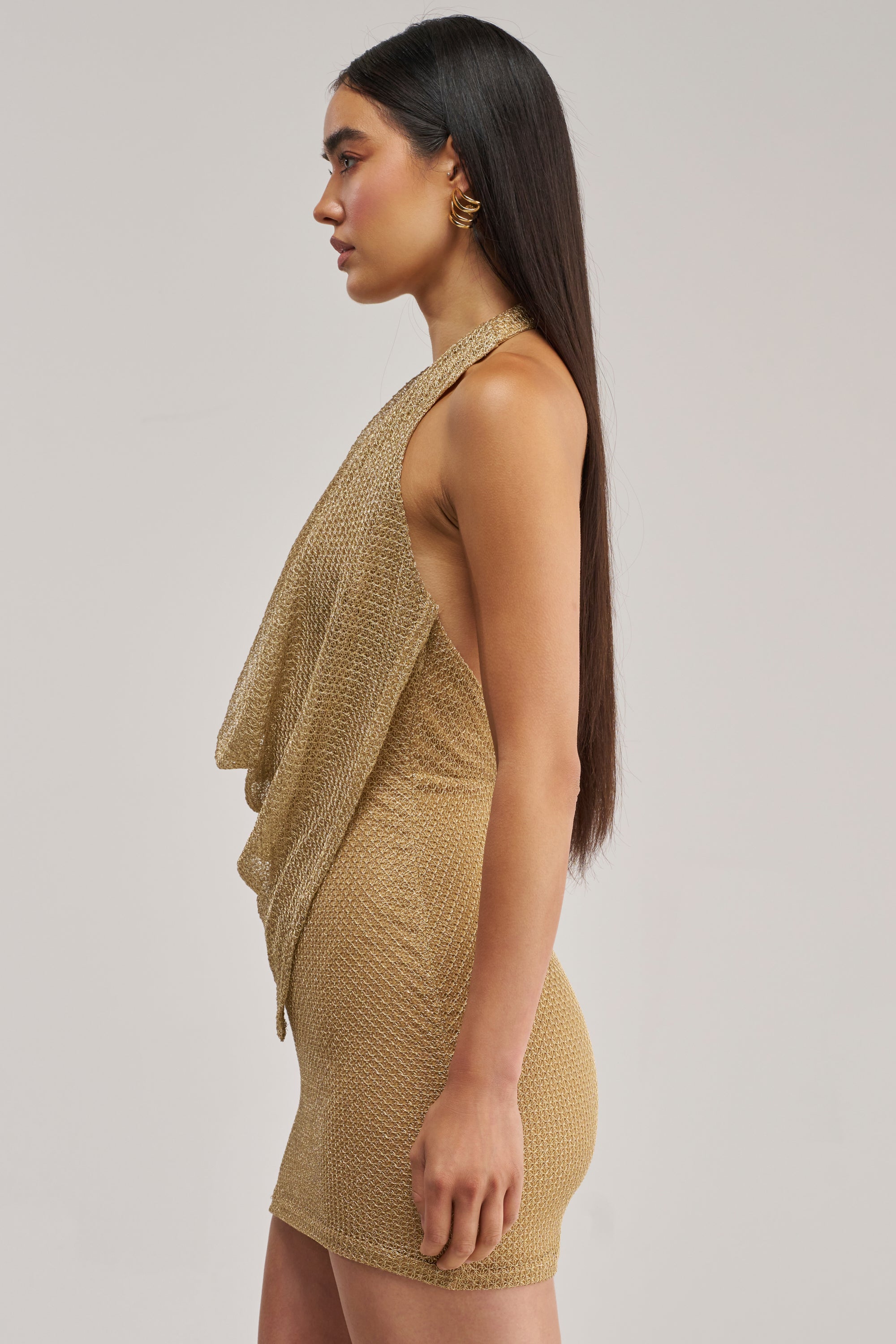 A woman with long, straight dark hair wears the ELECTRIC GLOW WOVEN MINI DRESS—a sleeveless, gold textured mini dress featuring a draped front—while standing in profile against a plain light background.