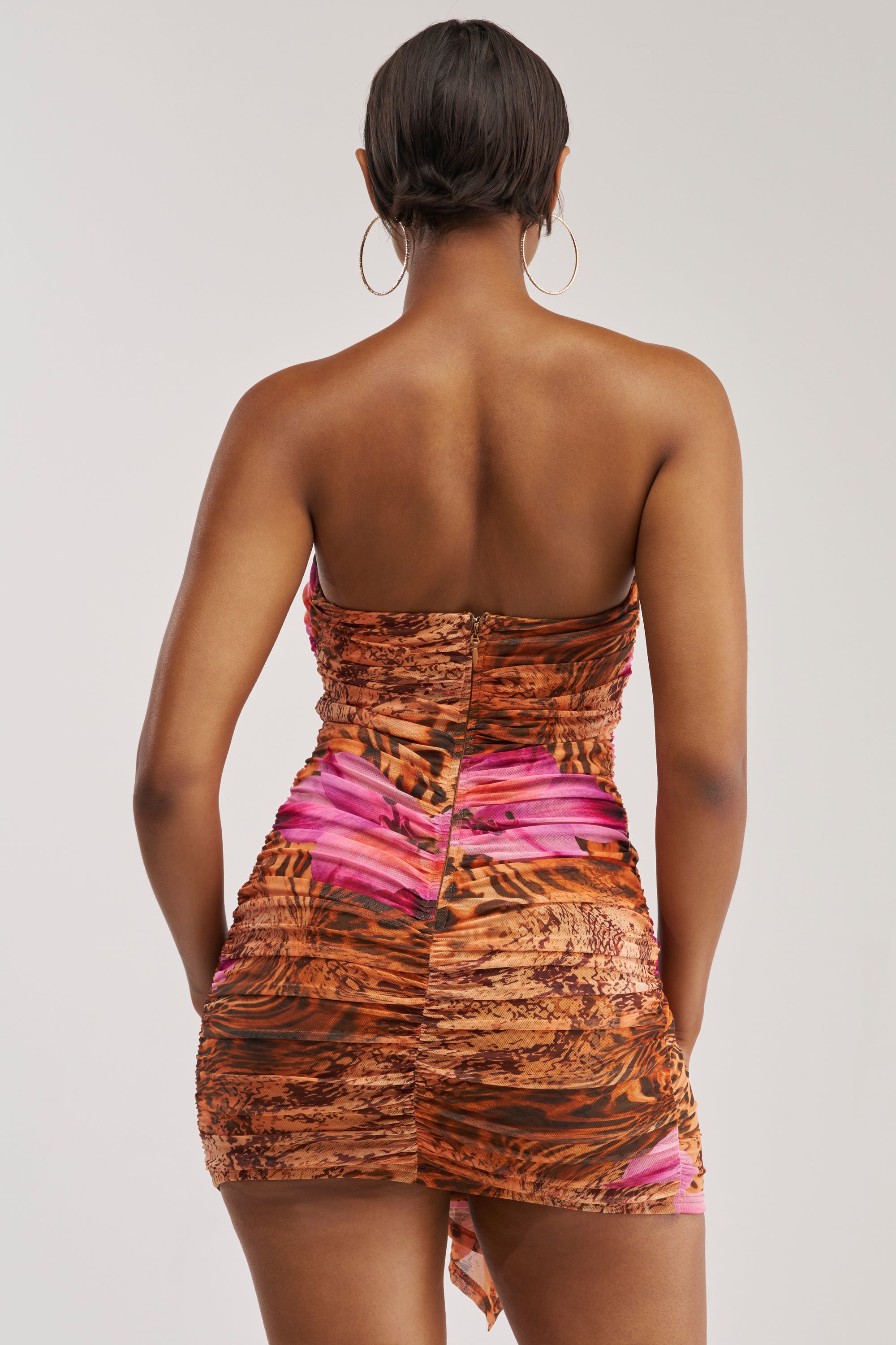 Wearing the BADDIE CLUB Plunging Mini Dress in a vibrant purple multi print, a person with short hair stands with their back to the camera against a plain background.