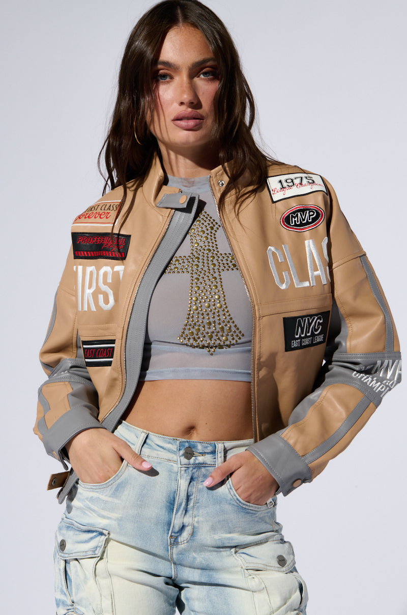 A person with long hair wears the FIRST CLASS EMBROIDERED MOTO jacket in beige, embellished with various patches, and pairs it with a gray crop top featuring an intricate beaded design. They complement the look with light-wash jeans, one hand tucked casually into a pocket, as they pose against a plain gray background.