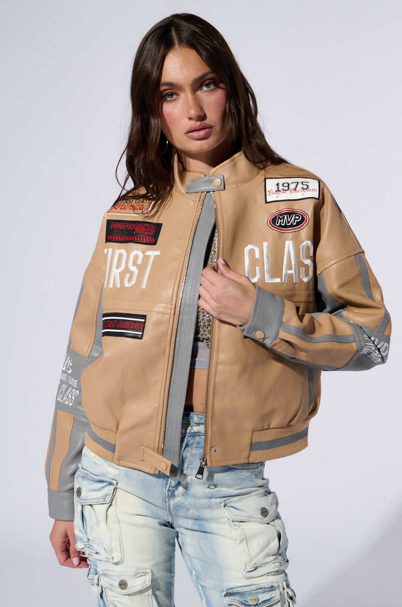 A woman with long, brown hair poses confidently in the First Class Embroidered Moto jacket, adorned with various patches and text, including the phrase "First Class." She pairs this faux leather outerwear with distressed light blue jeans, standing against a neutral background.