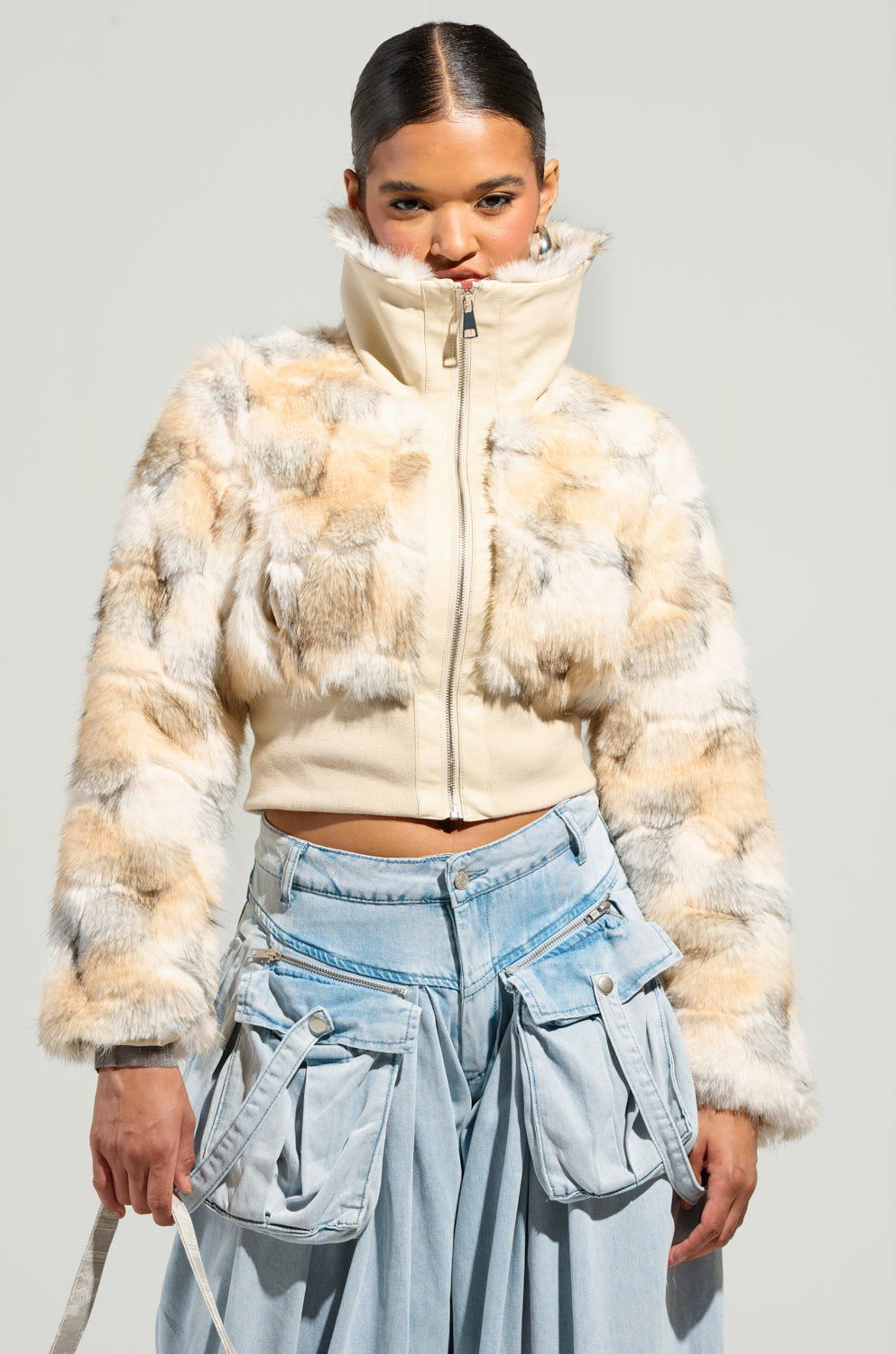 A person stands confidently in the SEE YOU AGAIN FITTED FUR BOMBER, characterized by its high collar, and pairs it with light blue, wide-legged cargo pants. The plain background accentuates their fashionable attire and pose.