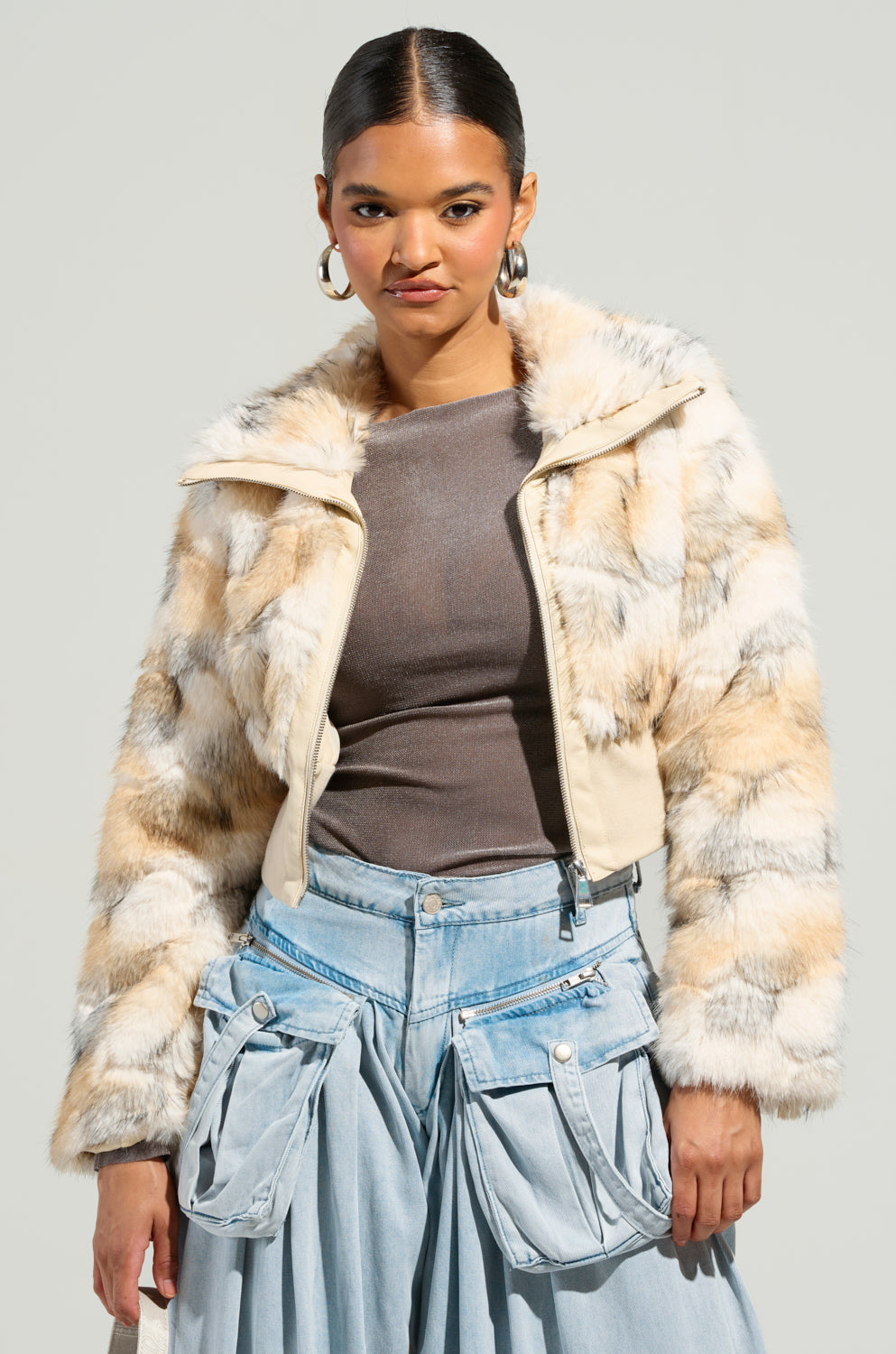 Wearing the SEE YOU AGAIN FITTED FUR BOMBER, an individual exudes confidence with its blend of beige, white, and brown tones over a brown top and light blue denim pants, complemented flawlessly by hoop earrings and sleek styled-back hair.
