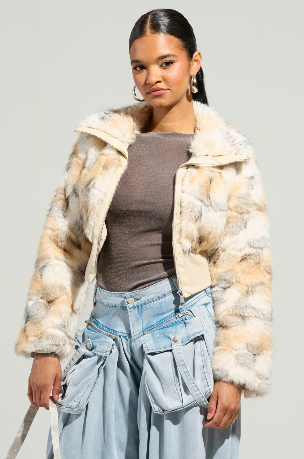 Dressed in the SEE YOU AGAIN FITTED FUR BOMBER, a trendy multi-toned faux fur jacket, over a brown top, the individual pairs it perfectly with wide-leg denim jeans that boast large front pockets. Completing the look are hoop earrings and dark hair sleekly styled back against a simple neutral backdrop.