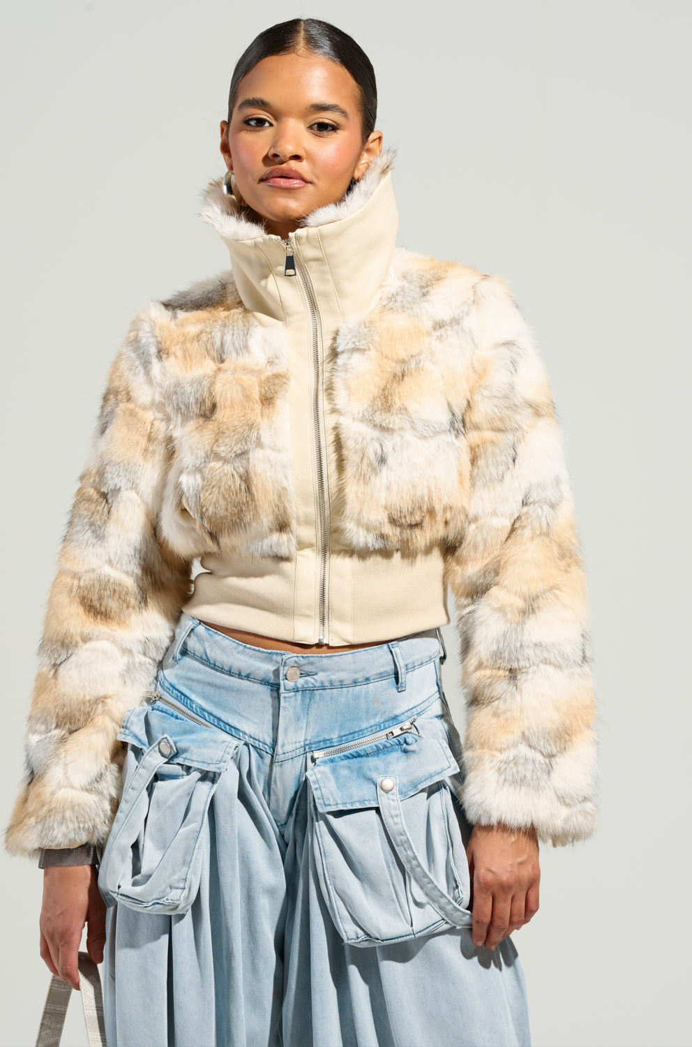 Wearing the SEE YOU AGAIN FITTED FUR BOMBER, its faux fur pattern enhancing the stylish cropped design, paired with high-waisted light blue denim pants featuring large pockets. The jacket is zipped up to the collar against an off-white background.