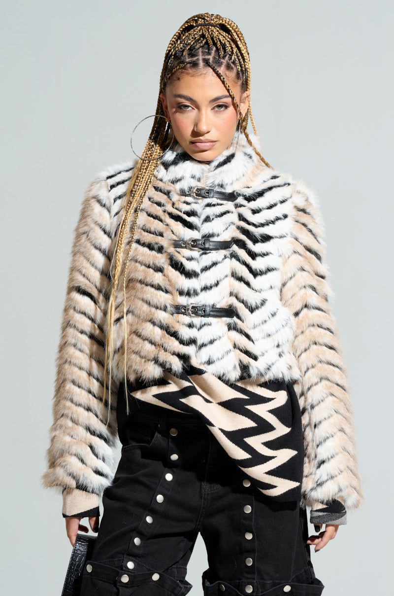 A person with braided hair strikes a confident pose, wearing the WILDER BUCKLE DETAIL FUR BOMBER featuring a tri-tone black and white striped pattern. Their black pants are accentuated with Wilder buckle detailing and adorned with buttons. Hoop earrings complete the ensemble against a neutral background.