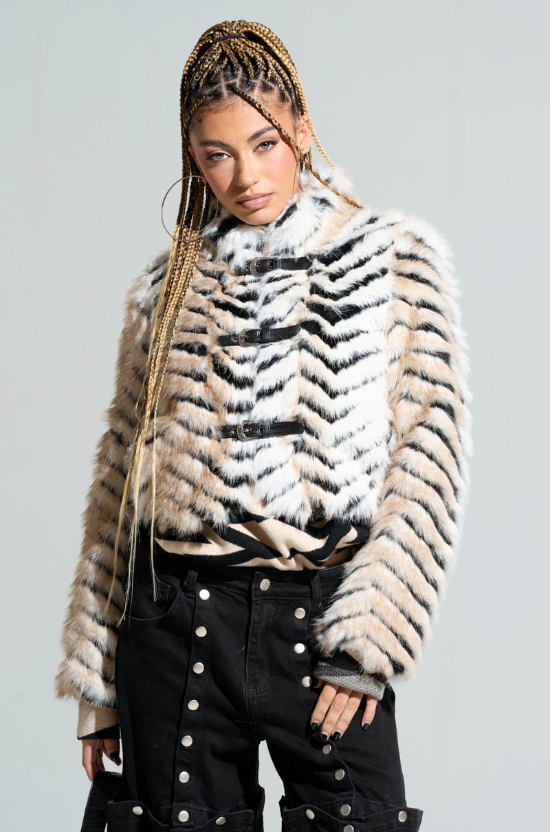 A person with long braided hair poses confidently in the tri-tone "Wilder Buckle Detail Fur Bomber," featuring a stylish black and white striped faux fur design. They are paired with high-waisted black pants, standing against a plain background.