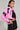 A person exudes confidence in a vibrant outfit featuring the BUBBLE POP ELECTRIC RACER MOTO faux leather jacket in bright pink and black, paired with a black crop top. Their short hair and earrings accentuate the moto-style skirt with lace-up detailing, while the plain background helps their striking ensemble stand out.