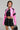 A person dressed in the vibrant BUBBLE POP ELECTRIC RACER MOTO jacket over a black top featuring text. They have dark hair and carry a chic faux leather handbag, completing their outfit with a black skirt that boasts a distinct crisscross pattern, adding an edgy touch to their colorful look.