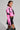 A woman strikes a confident pose in the BUBBLE POP ELECTRIC RACER MOTO, a bright pink and black moto-style jacket with a matching mini skirt featuring lace-up details. Her faux leather handbag, adorned with spikes, complements her bold earrings against the plain gray backdrop.