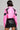 From behind, a person with short hair is wearing the BUBBLE POP ELECTRIC RACER MOTO, a bright pink and black moto-style cropped faux leather jacket. It's paired perfectly with a matching black leather mini skirt featuring lace-up details on the side. Their tattoos are visible on their legs, enhancing the Bubble Pop Electric vibe.