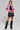A person stands confidently in the BUBBLE POP ELECTRIC RACER MOTO bright pink jacket, paired with a black crop top and ruffled black mini skirt. They sport tall black boots and carry a small faux leather handbag. With short hair against a plain gray background, their look exudes edgy sophistication.