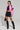 A person poses confidently, wearing a bright pink cropped jacket and a black lace-up mini skirt. They sport knee-high BUBBLE POP ELECTRIC RACER MOTO boots and hold a small studded bag. Their hair is short and slicked back, and they have visible tattoos on their legs.