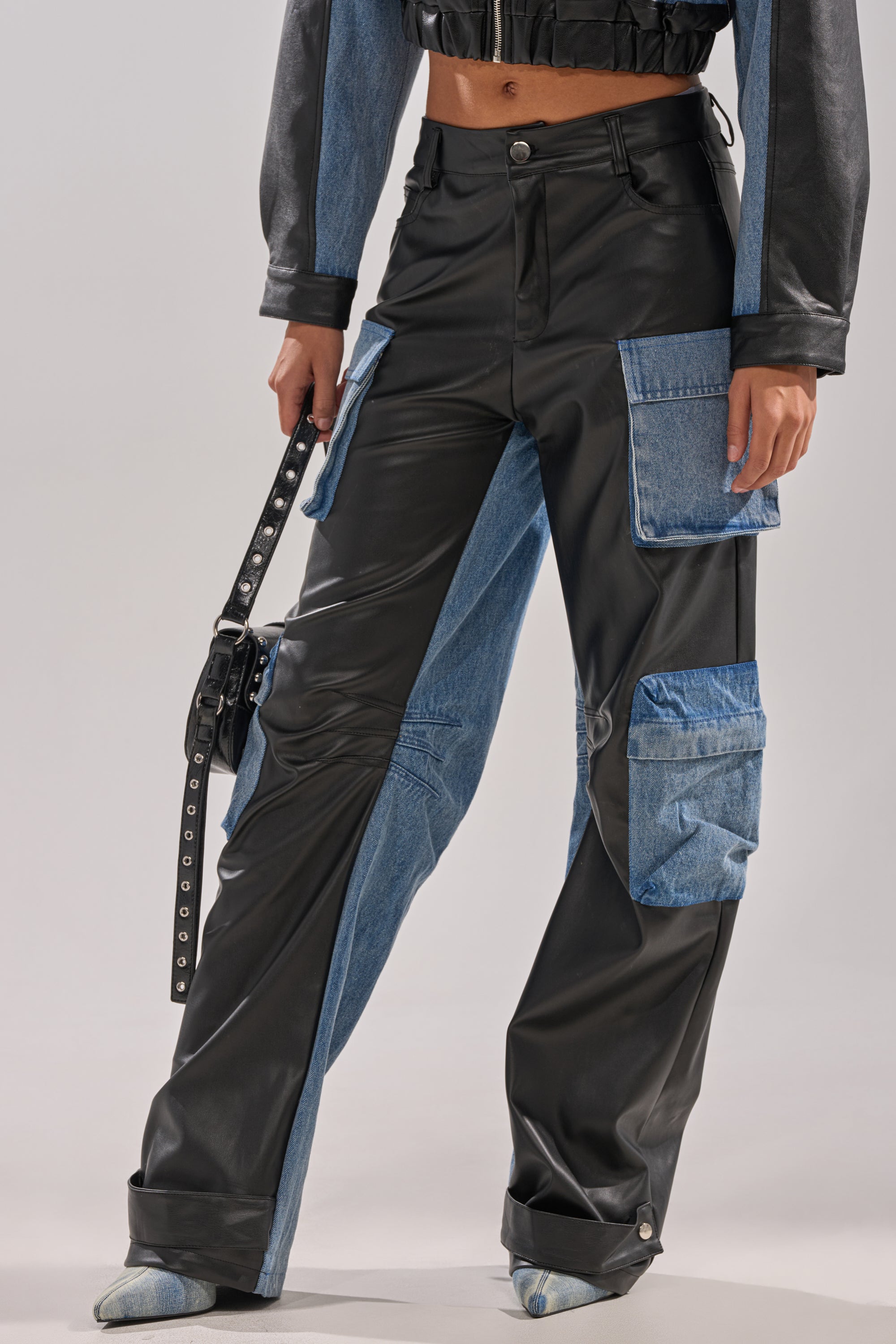 A person wears the DOWN TIME MIXED MEDIA CARGO PANT, featuring high-waisted faux leather with blue denim patches and large utility pockets, paired with a matching black cropped jacket and a studded black belt.