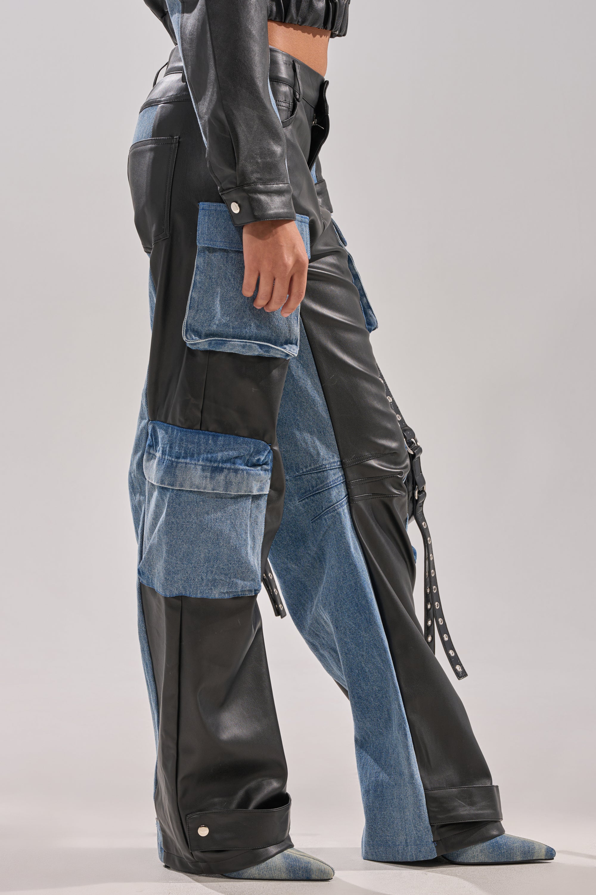 A person wears the DOWN TIME MIXED MEDIA CARGO PANT—high-waisted, wide-leg cargo pants with black faux leather and blue denim patchwork, large utility pockets, ankle snap buttons—paired with a cropped black leather jacket.