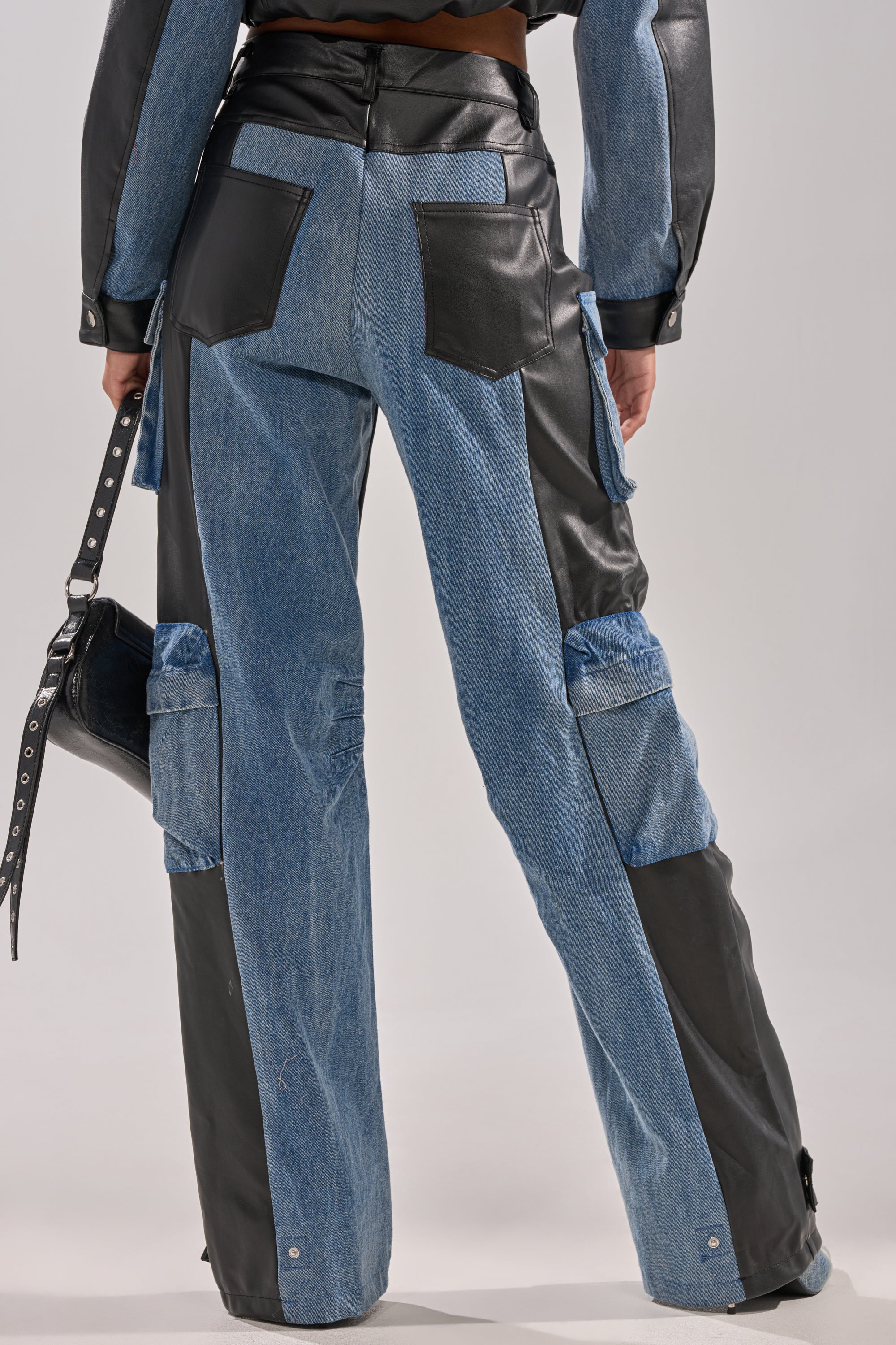 A person stands facing away, wearing the DOWN TIME MIXED MEDIA CARGO PANT—utility-style denim and black faux leather cargo pants with cargo pockets, black back pockets, and holding a matching bag by their side.