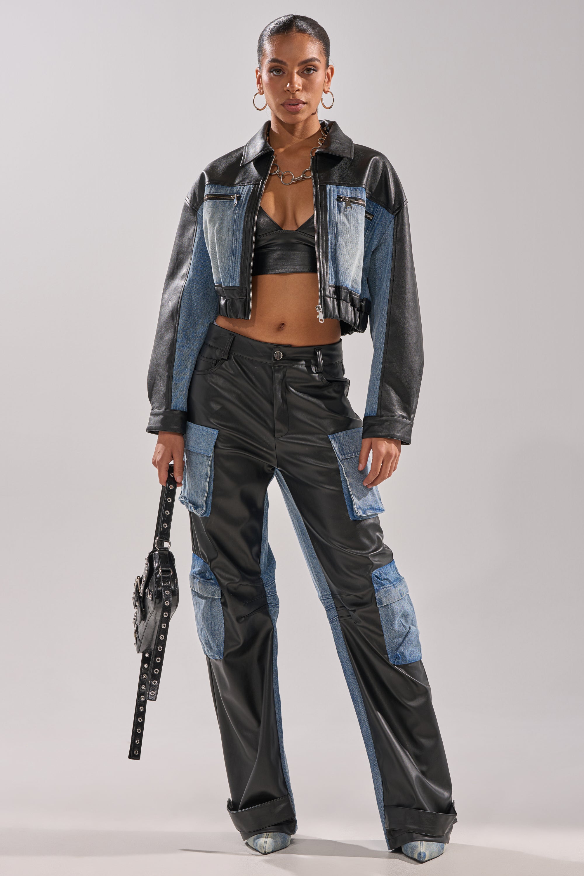 A woman stands confidently in DOWN TIME MIXED MEDIA CARGO PANT—a black faux leather and denim patchwork pant—paired with a matching jacket, black crop top, hoop earrings, and studded black handbag against a plain light gray background.