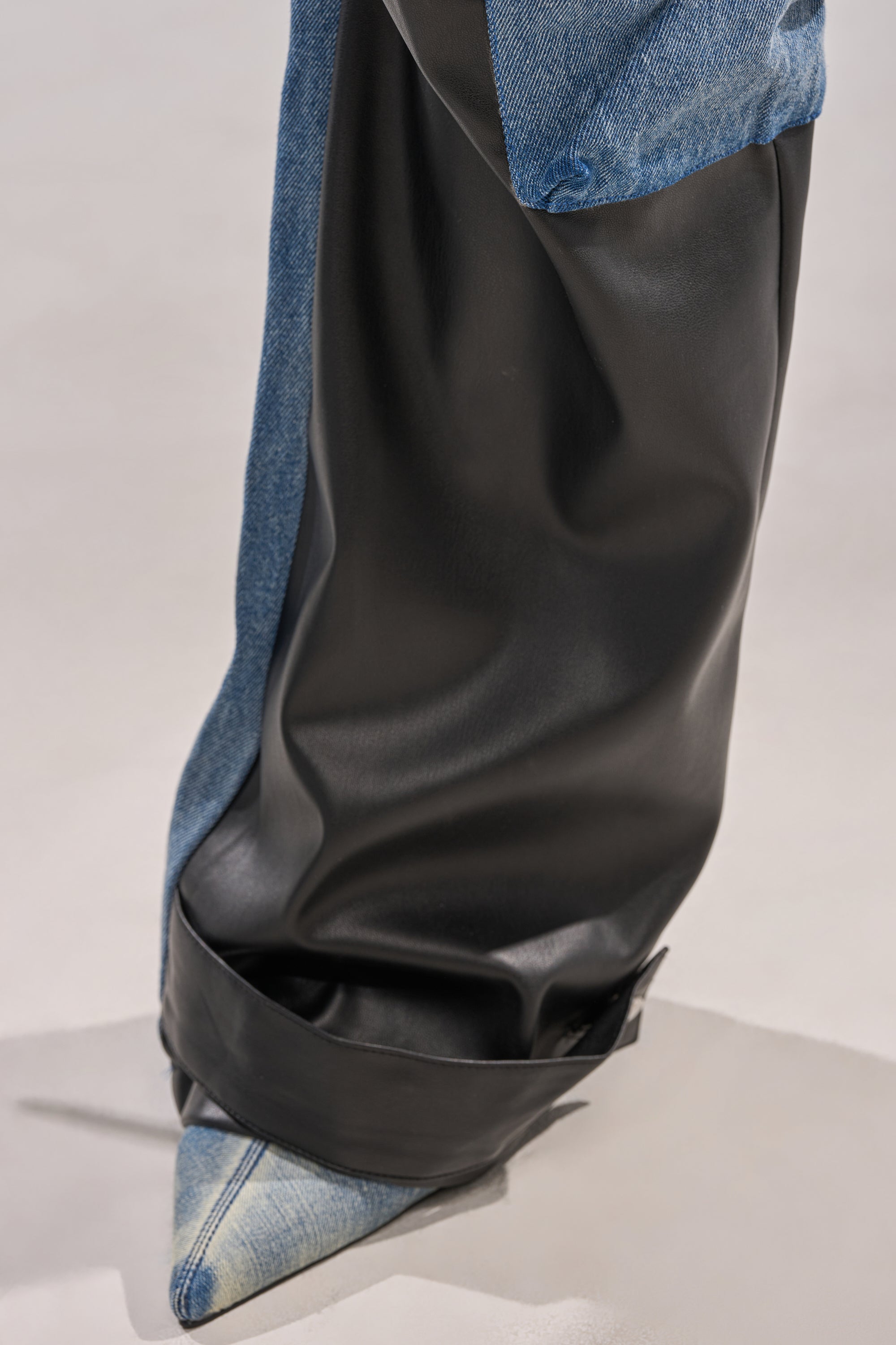 A close-up of a person in the DOWN TIME MIXED MEDIA CARGO PANT featuring one denim leg and one black faux leather leg over pointed-toe denim shoes, with the pants covering most of the shoes against a light background.