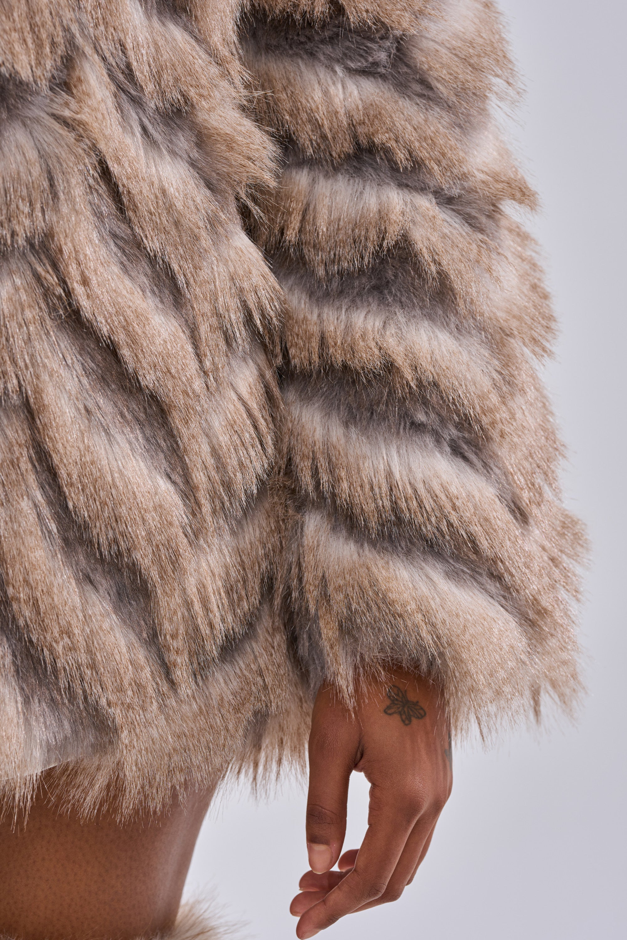 A close-up of an arm in the SHOWBIZ BABY FAUX FUR coat, featuring a chevron print faux fur in tan, brown, and gray. A tattooed hand peeks out just below the cozy sleeve.