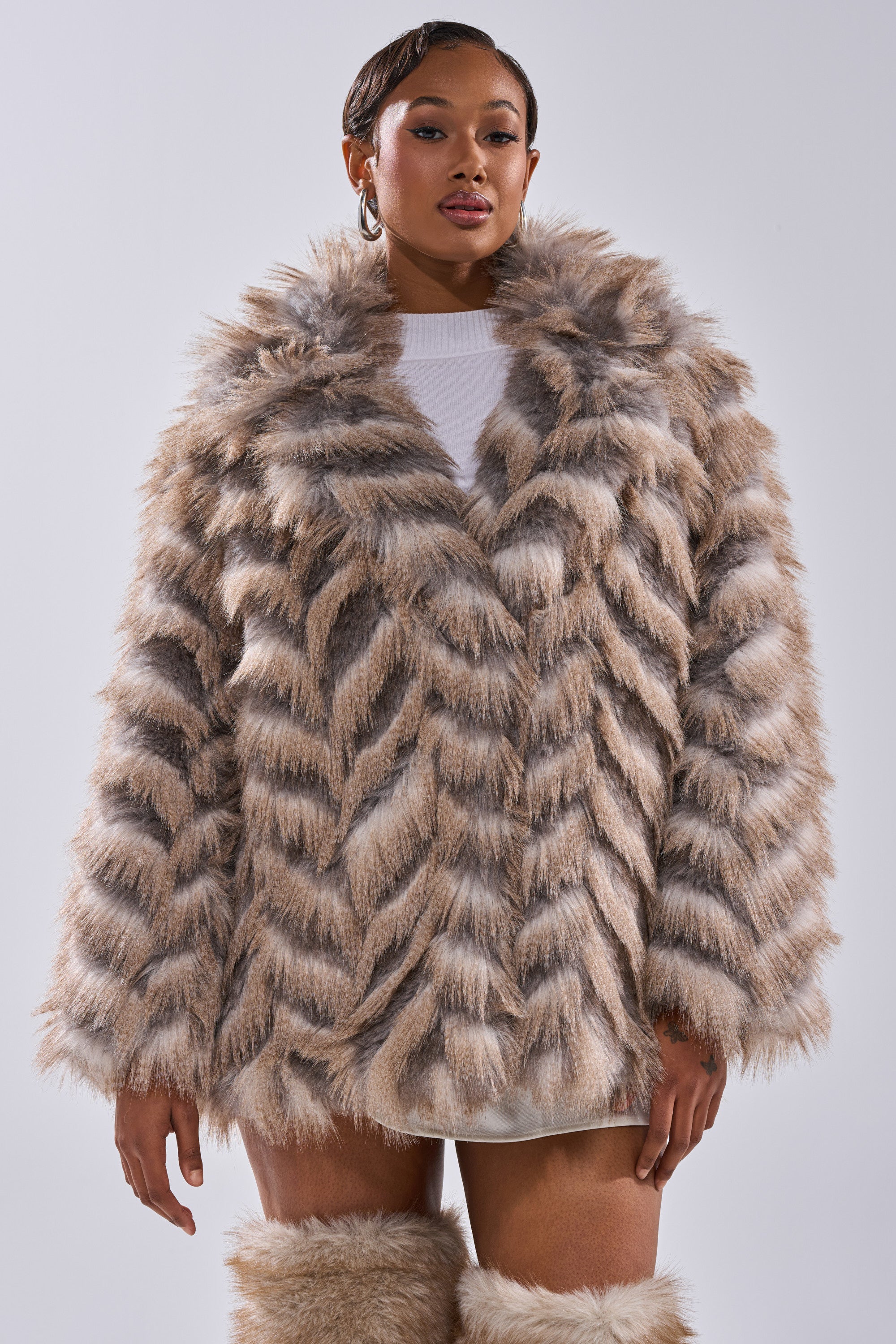 A woman poses confidently in the SHOWBIZ BABY FAUX FUR longline chevron coat, styled with hoop earrings, a white top, and matching faux fur boots, against a plain light background.